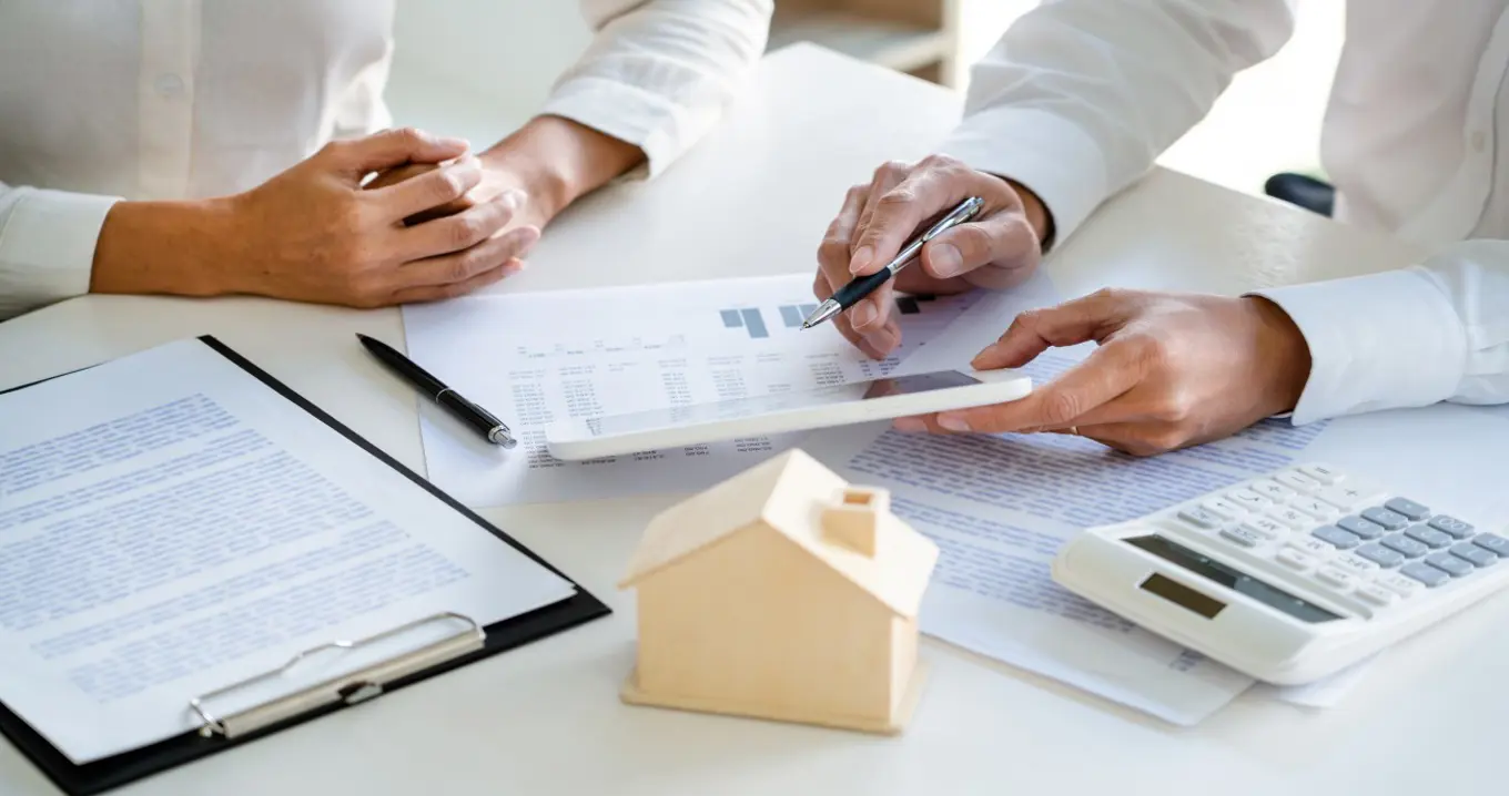 Business professionals reviewing financial documents and real estate data at a desk, with a small model house and calculator symbolizing property investment planning.