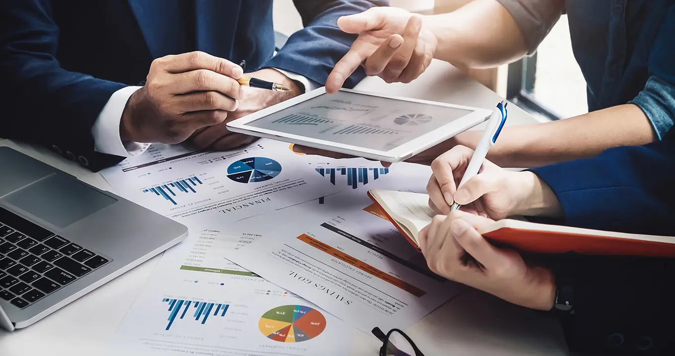 Close-up of business professionals analyzing financial charts and graphs on paper and tablet during a strategic planning meeting, surrounded by documents and a laptop.
