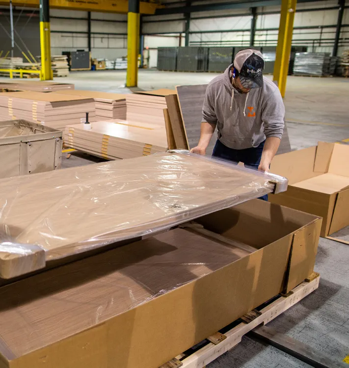 Worker in a warehouse handling a large flat wooden board wrapped in plastic over an open cardboard box on a pallet