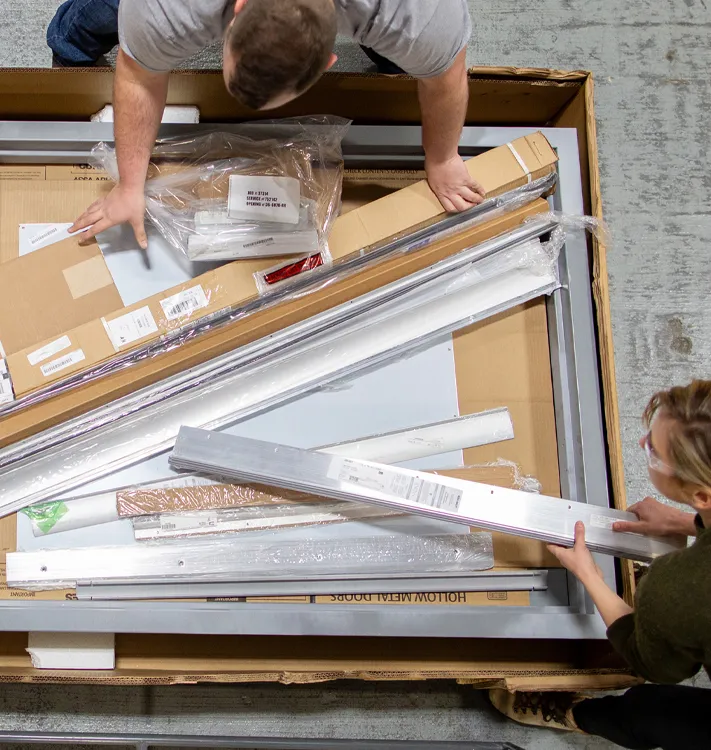 Two people unpacking door and lock components from a large cardboard box on a warehouse floor