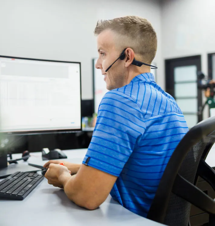 Man wearing a blue striped shirt and headset sitting at a desk, looking at a computer screen