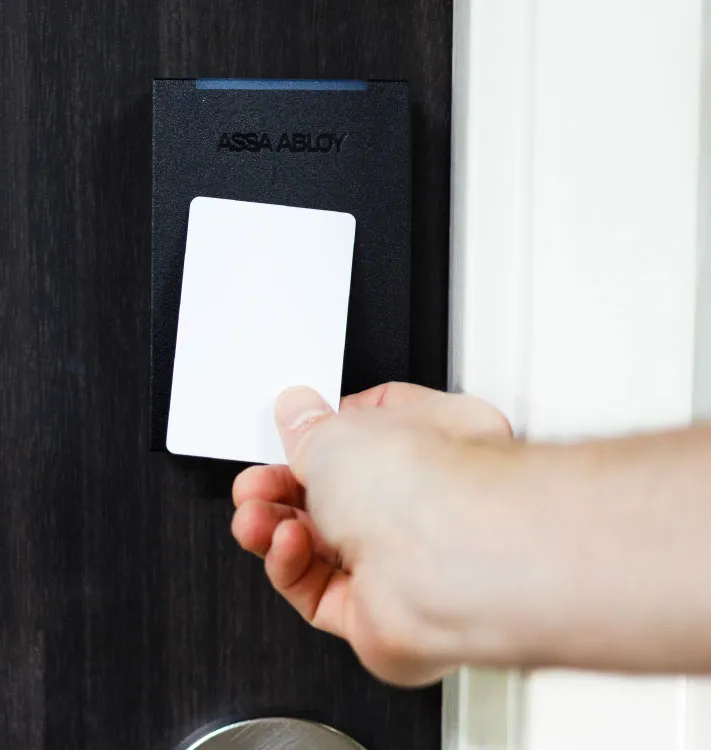 Hand holding a white card near a black ASSA ABLOY RFID card reader on a dark door