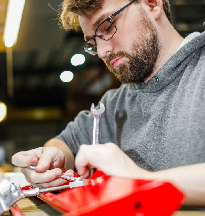 Man with glasses and beard focused on repairing a red mechanical device using a wrench.