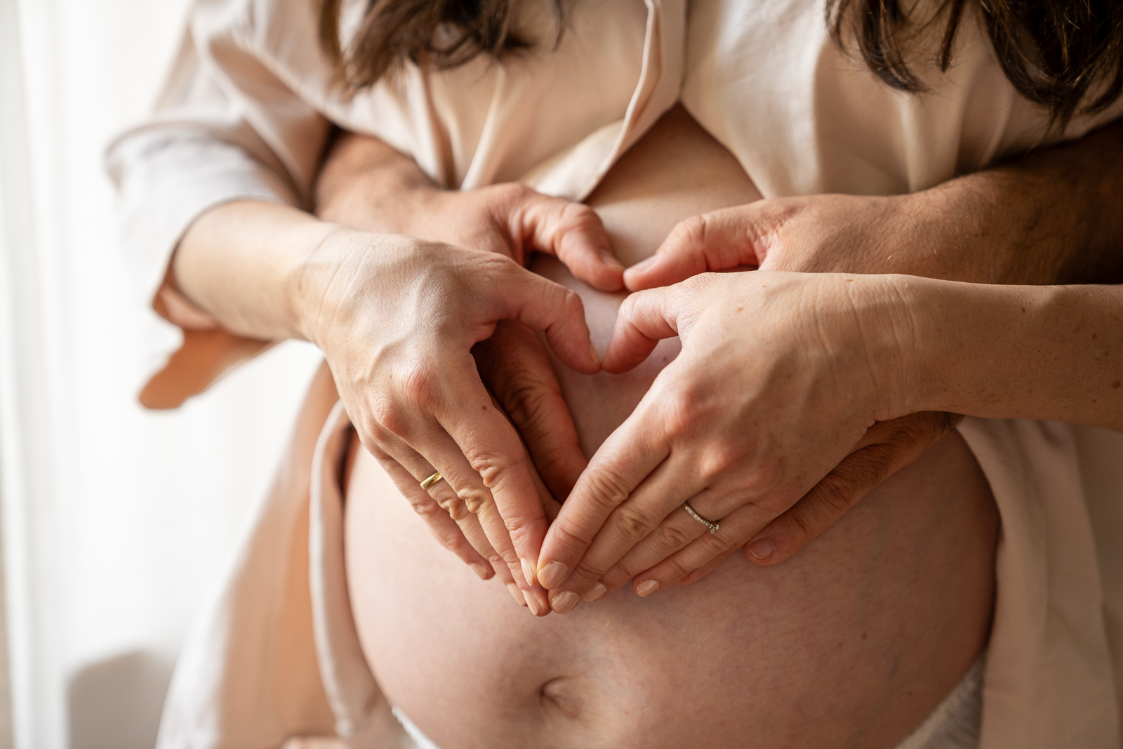 Babybauch Shooting Paarfoto – Hände in Herzform auf dem Bauch einer schwangeren Frau, liebevoller Moment festgehalten in natürlichem Licht