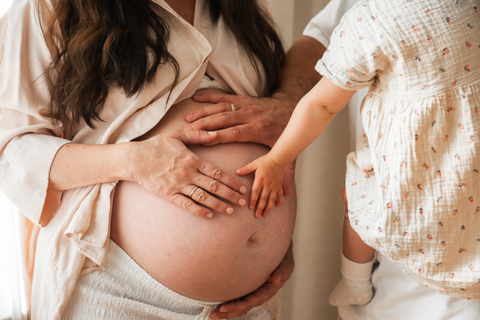 Intime Babybauchfotografie – werdende Eltern und Geschwisterkind legen die Hände liebevoll auf den schwangeren Bauch, festgehalten im natürlichen Licht.