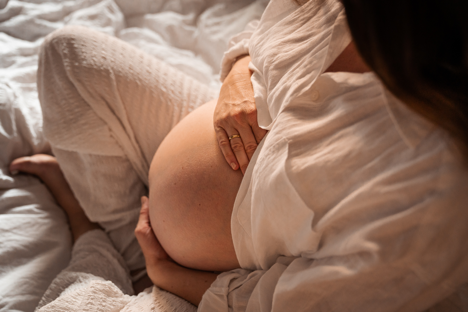 Babybauch Shooting zu Hause – Schwangere sitzt gemütlich auf dem Bett und hält liebevoll ihren Bauch, warme Lichtstimmung für authentische Schwangerschaftsfotos.