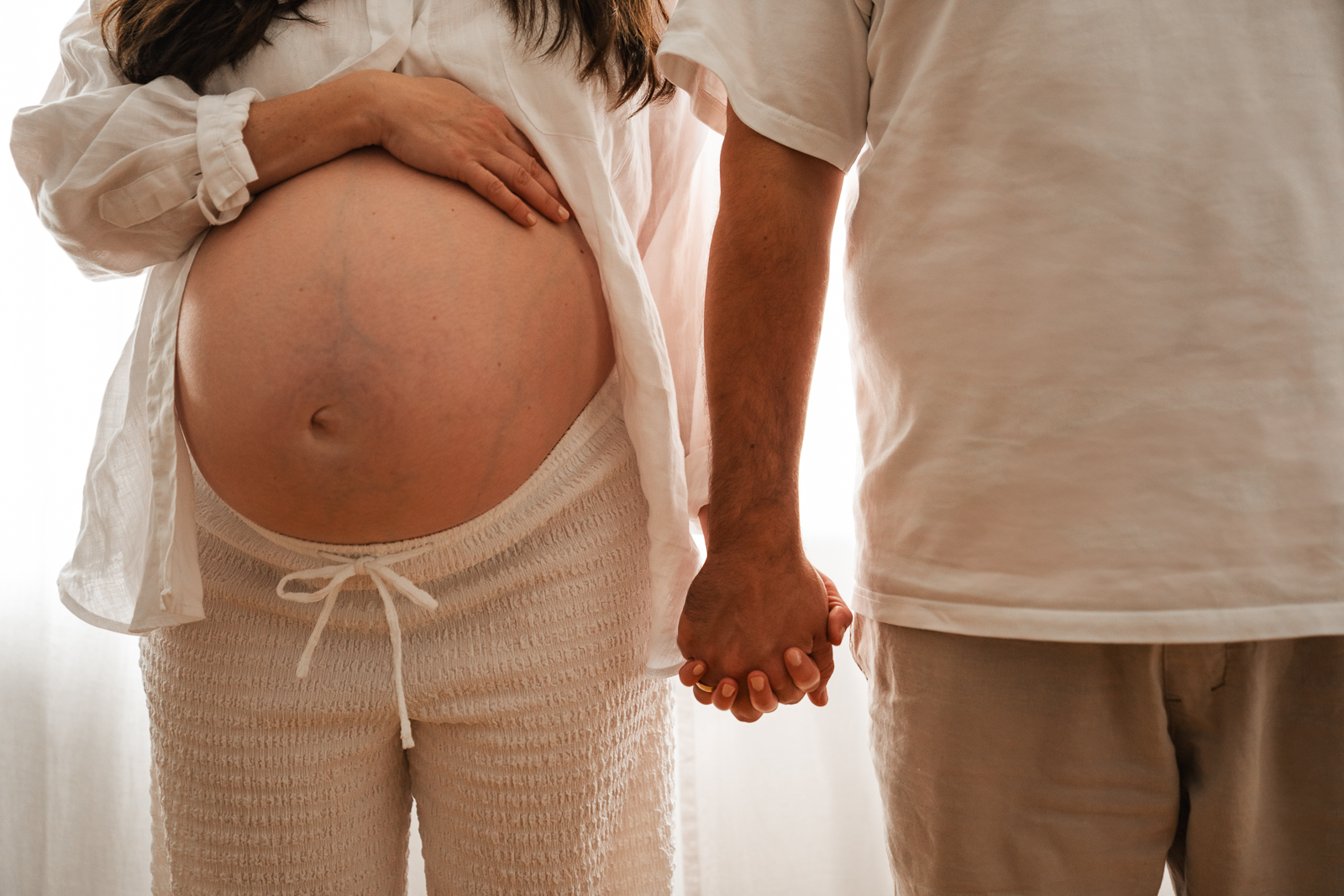 Babybauch Shooting Paarfoto – werdende Eltern halten sich an den Händen, Fokus auf den runden Bauch und harmonische Farbabstimmung in Weißtönen.