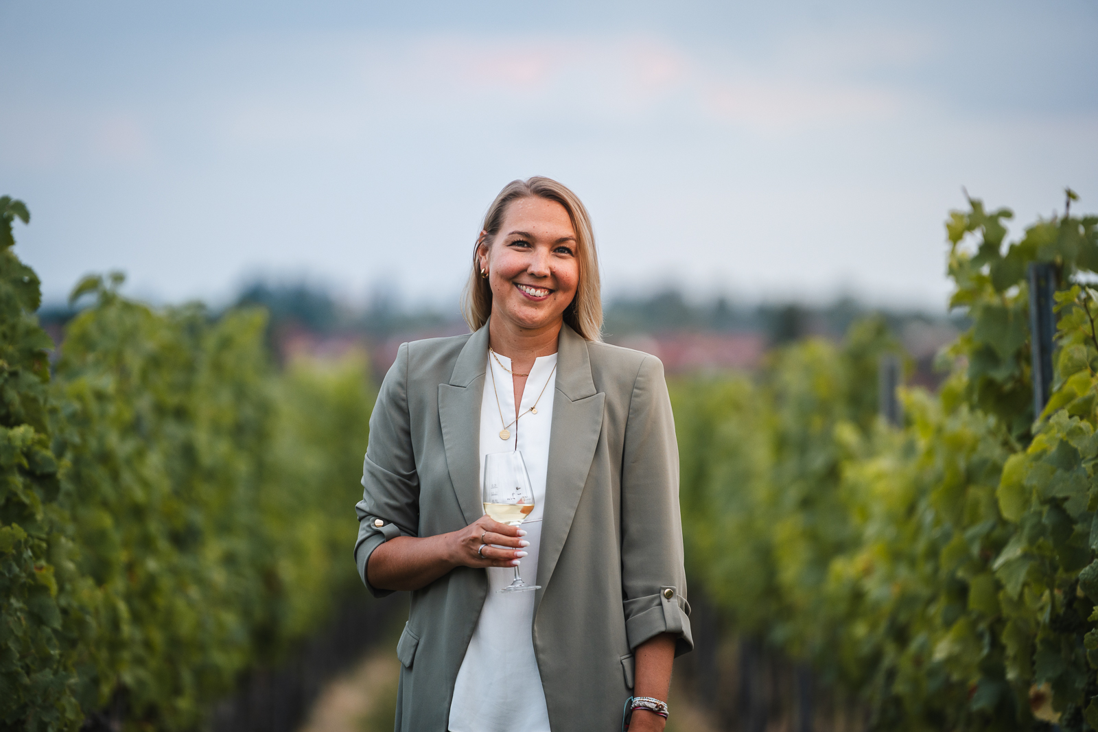 Frau im Blazer und weißem Oberteil steht mit einem Glas Weißwein im Weinberg und lächelt in die Kamera – professionelle Portraitfotografie
