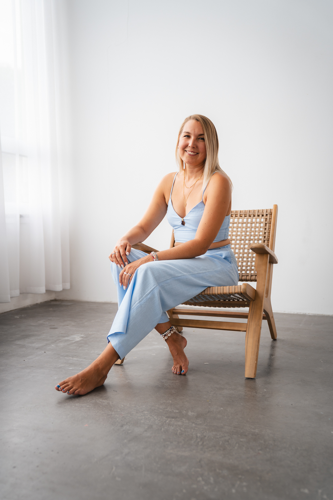 Frau in hellblauem Jumpsuit sitzt entspannt barfuß auf einem Stuhl im modernen Fotostudio – Lifestyle Portrait