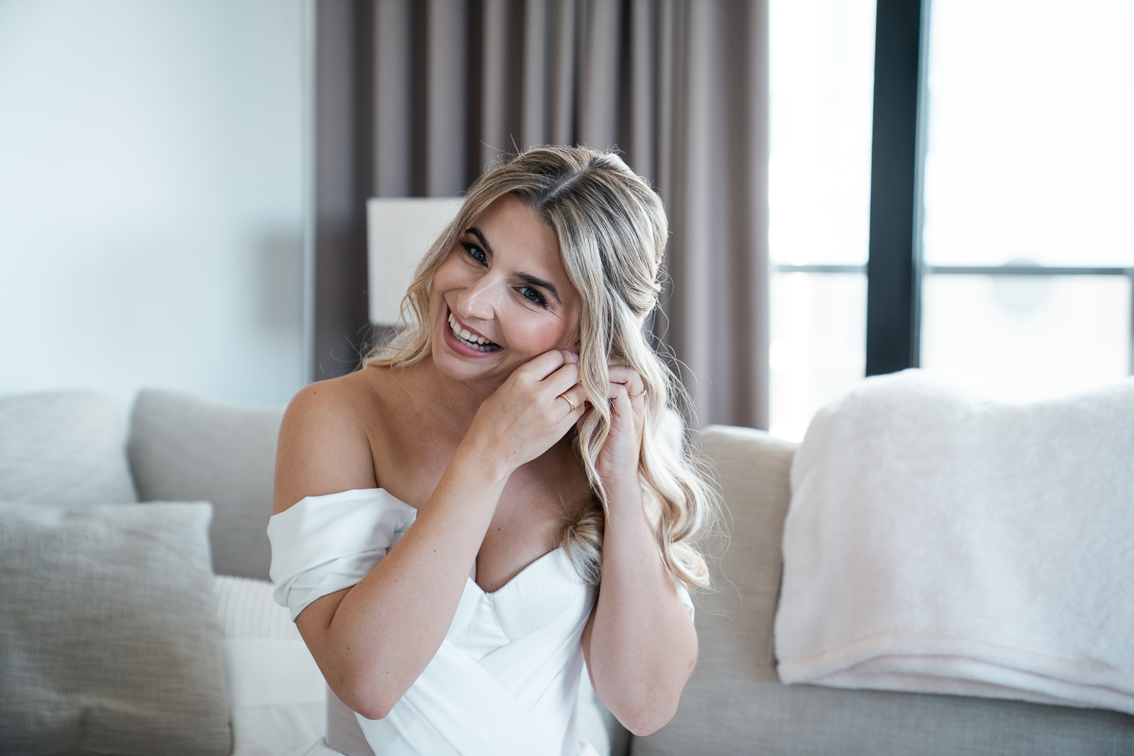 Braut im weißen Hochzeitskleid steckt Ohrringe an und lacht während des Getting Ready, natürliche Hochzeitsfotografie