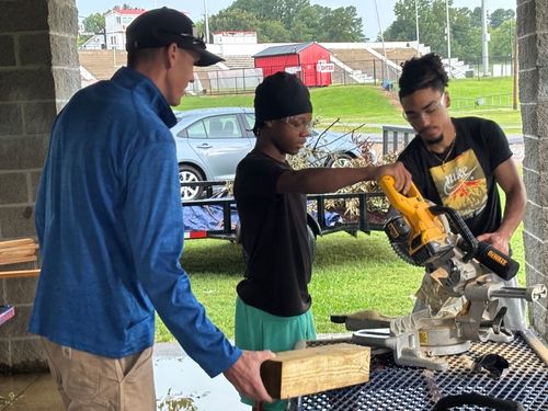 Two young volunteers in safety glasses cut lumber with a miter saw while an adult volunteer holds a board, working under a covered pavilion.