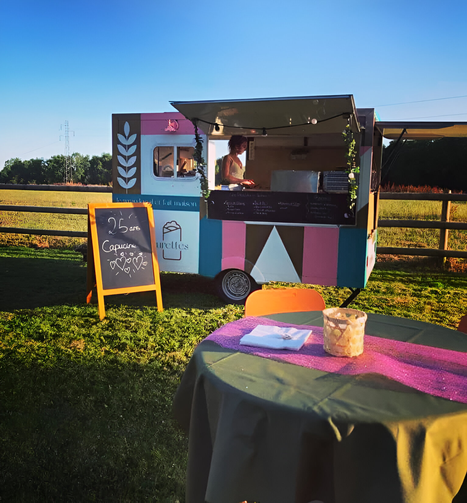 Camion de nourriture coloré avec une femme servant à l'intérieur, une ardoise indiquant «25 ans Capucine» et une table avec une nappe verte et un chemin de table rose à l'avant.