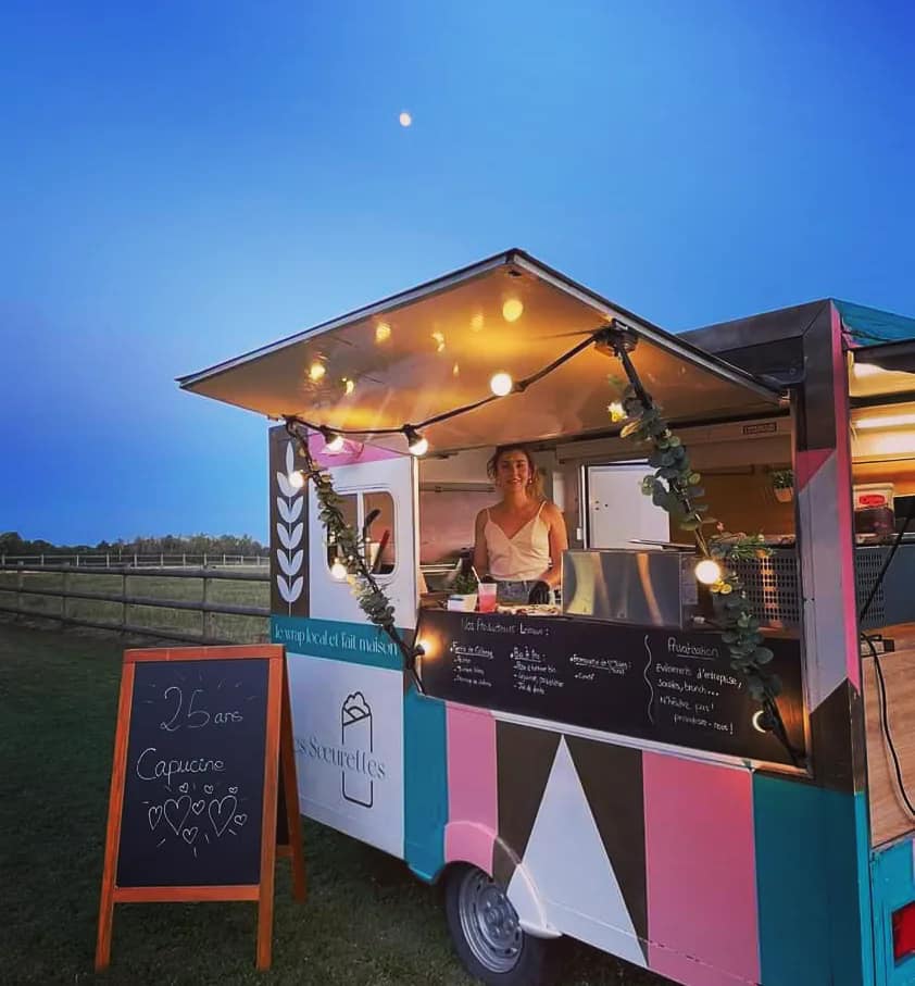 Une femme souriante dans un food truck coloré décoré de guirlandes lumineuses au crépuscule avec un panneau indiquant 25 ans Capucine.