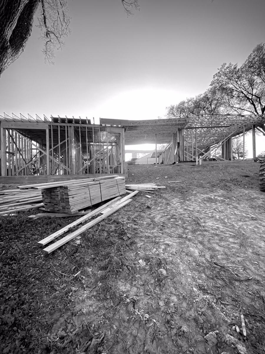 Construction site featuring wooden house framing with stacked lumber and surrounding trees.