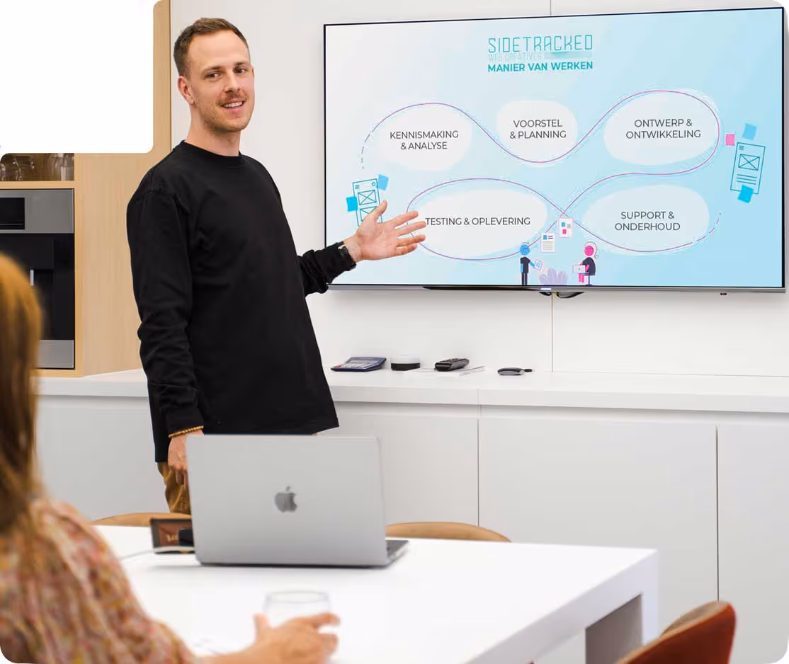 Man presenting a workflow diagram on a screen in a modern office, with a laptop and seated audience member visible.