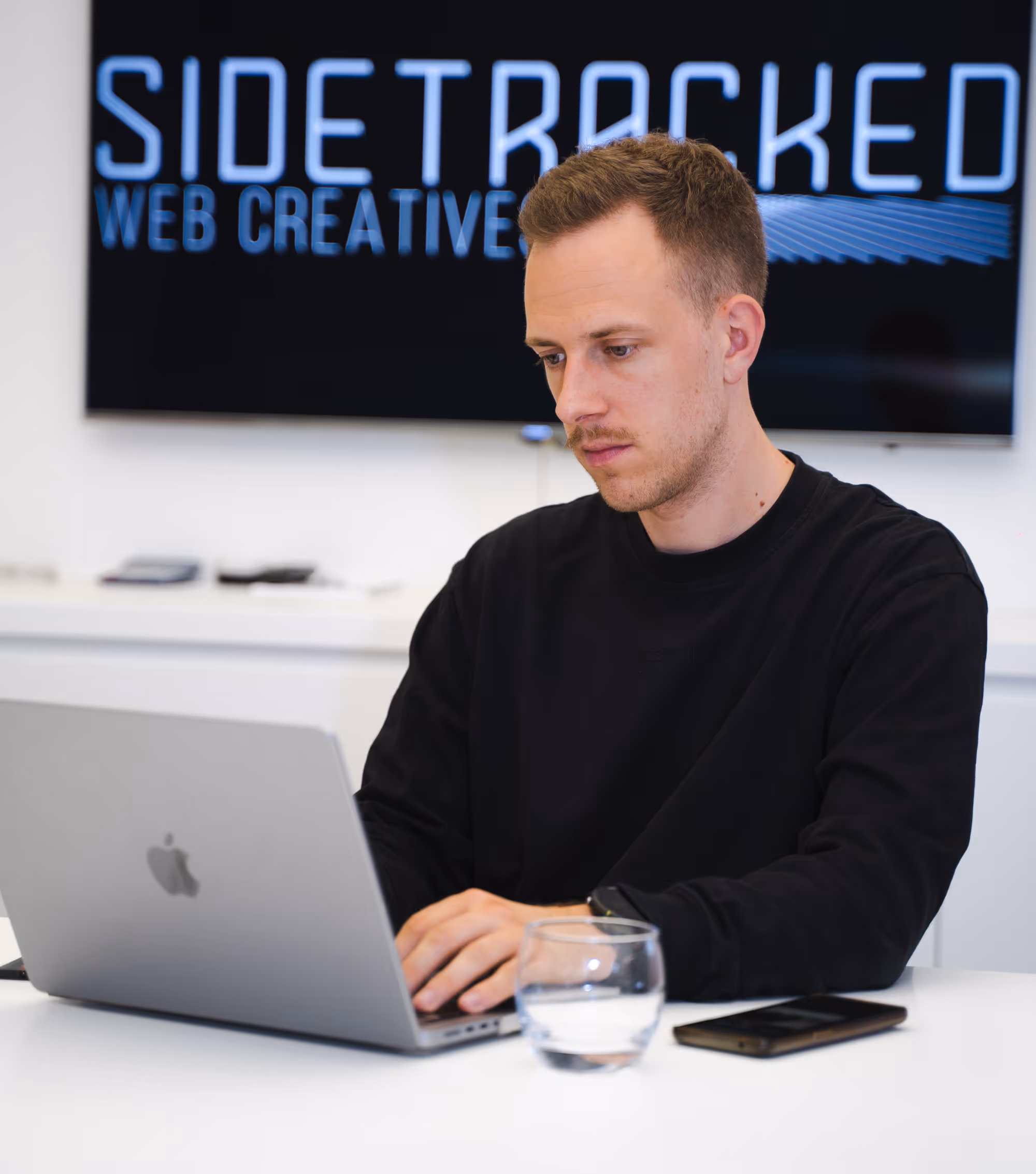 Man in black shirt working on a MacBook laptop at a white desk with a glass of water and a smartphone nearby, with a screen behind him displaying 'SIDETRACKED WEB CREATIVE'.