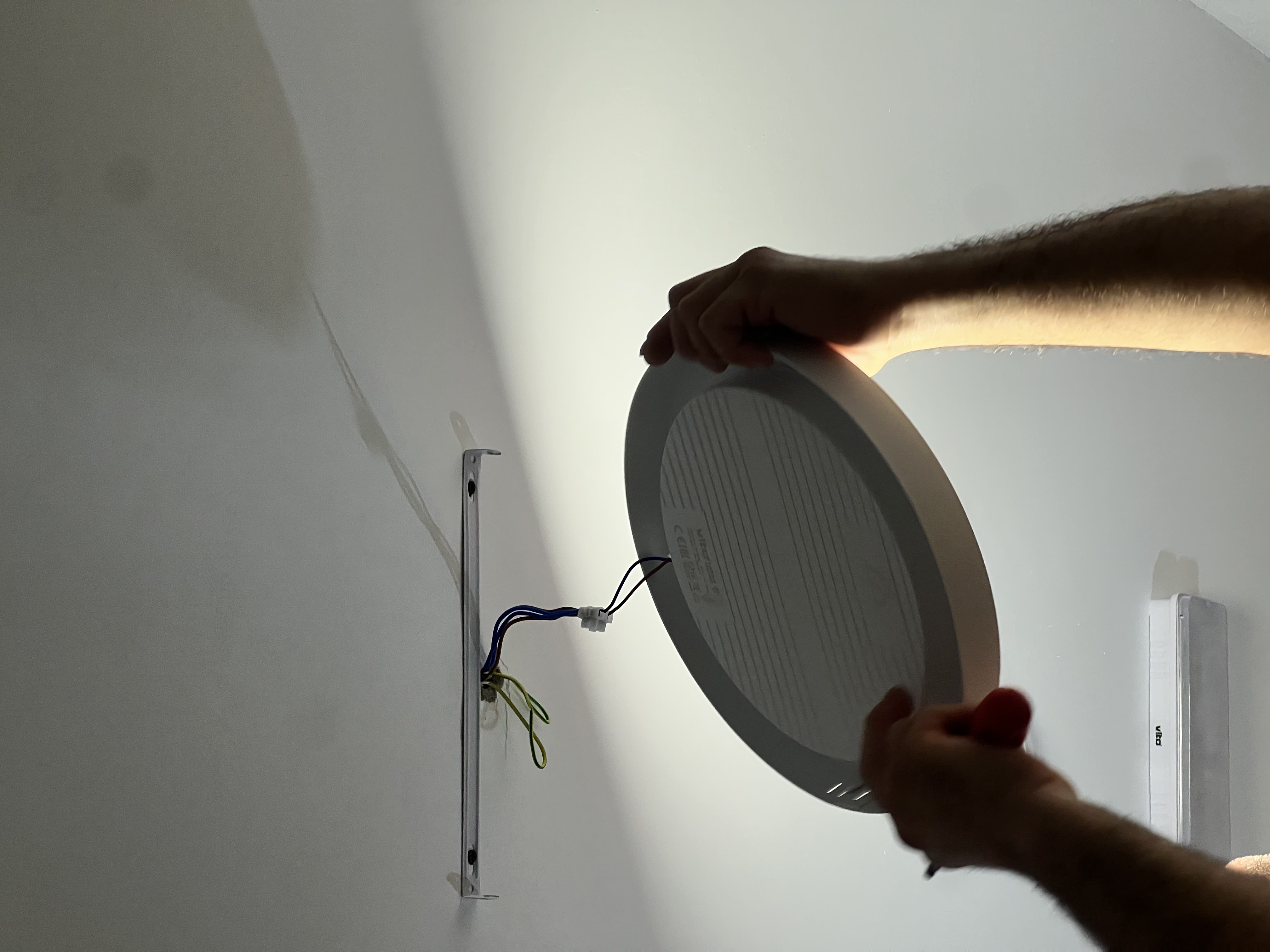 Person installing a round ceiling light fixture with exposed wiring on a white ceiling.
