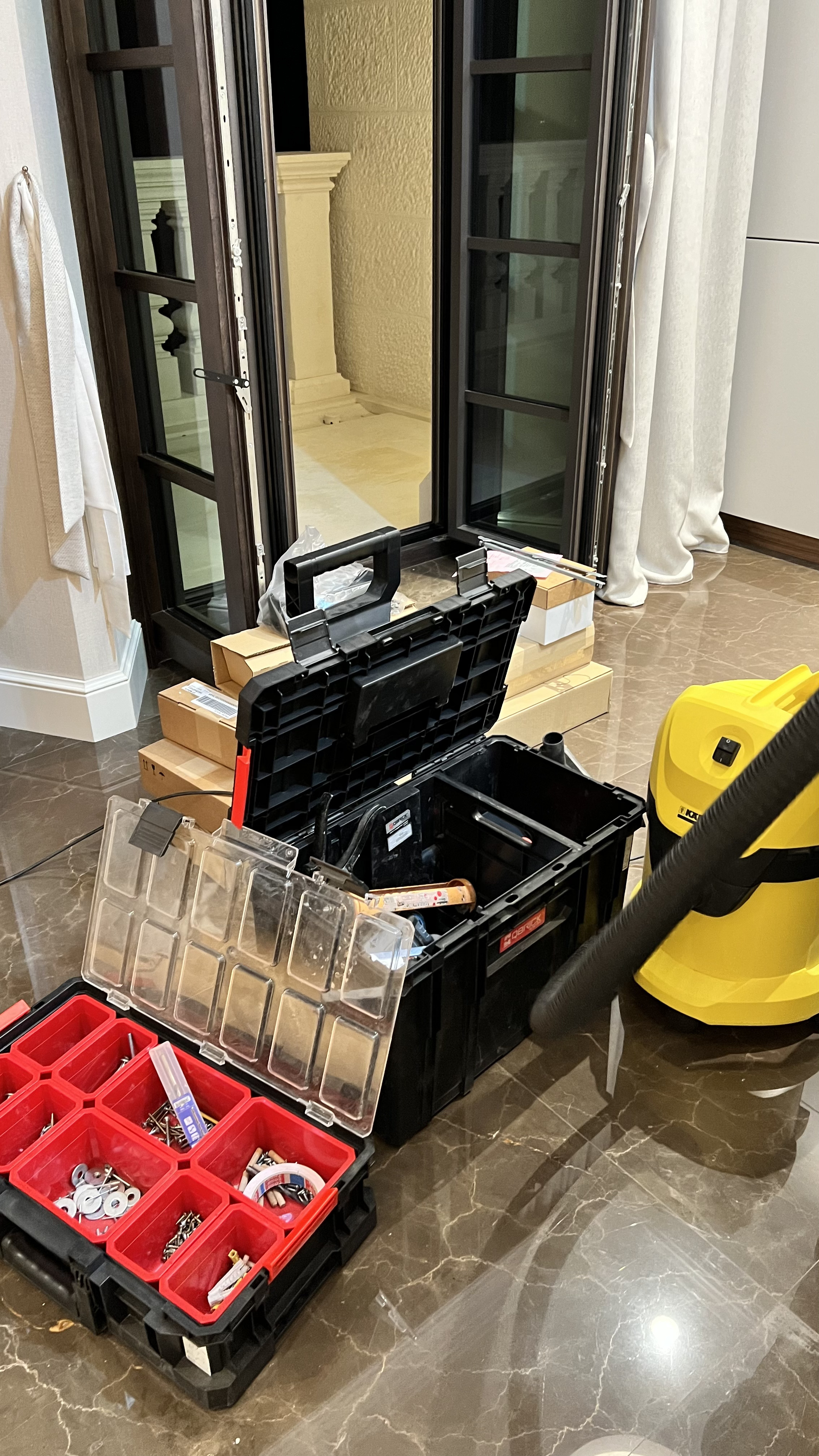 Open black toolboxes with screws, washers, and other small hardware on a polished floor next to a yellow vacuum cleaner and glass doors.