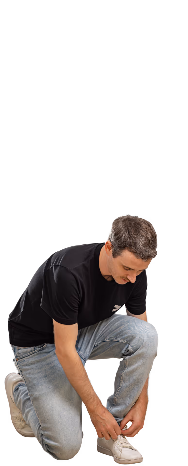 Ein Mann, Dominik Hutter, im schwarzen T-Shirt und Jeans kniet, um sich einen weissen Turnschuh zu binden.