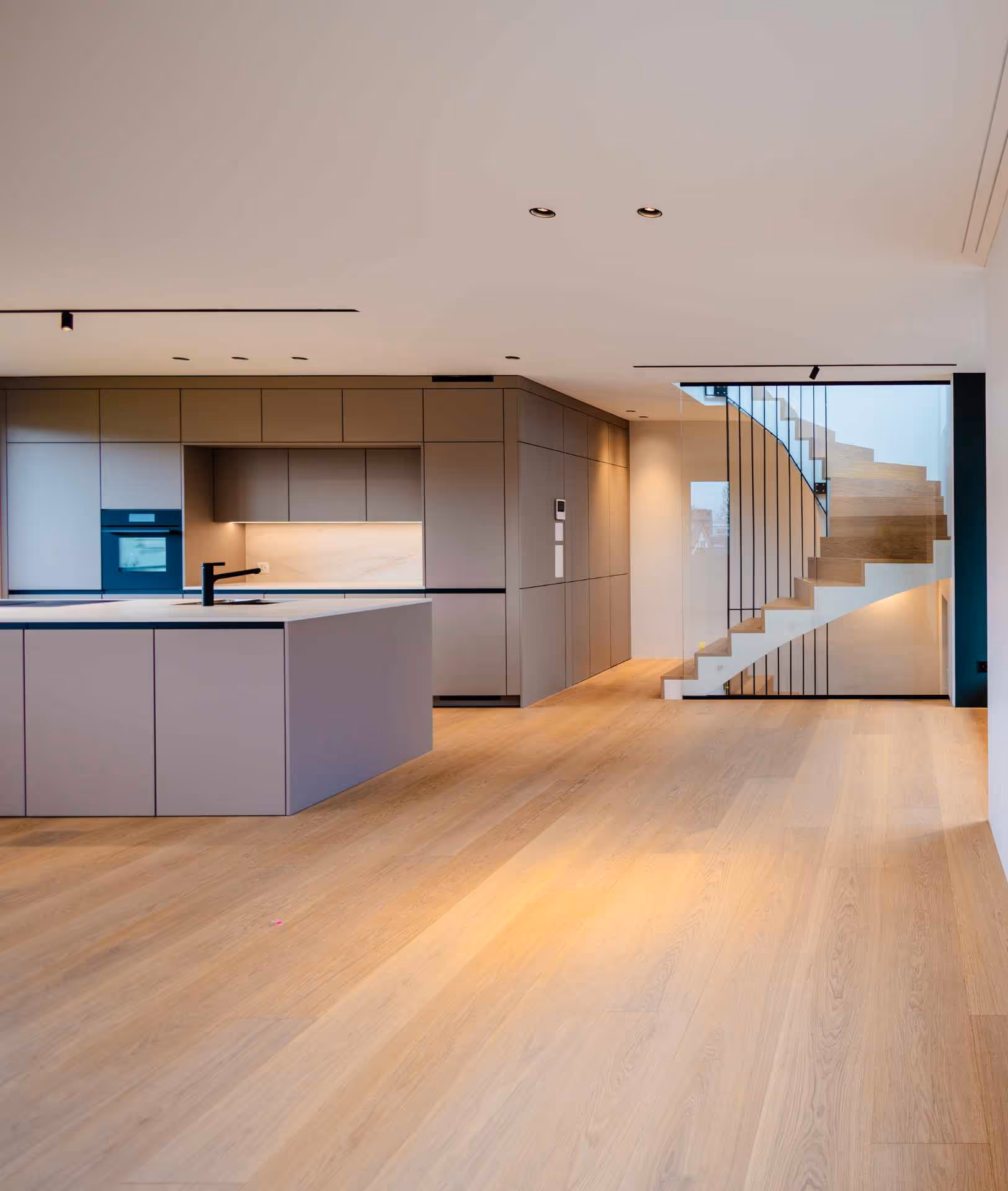 Moderne helle Küche mit Insel, grauen Schränken und Holztreppe mit Glasgeländer.