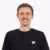 Junger Mann mit kurzen braunen Haaren und schwarzem Shirt lächelt vor weißem Hintergrund.