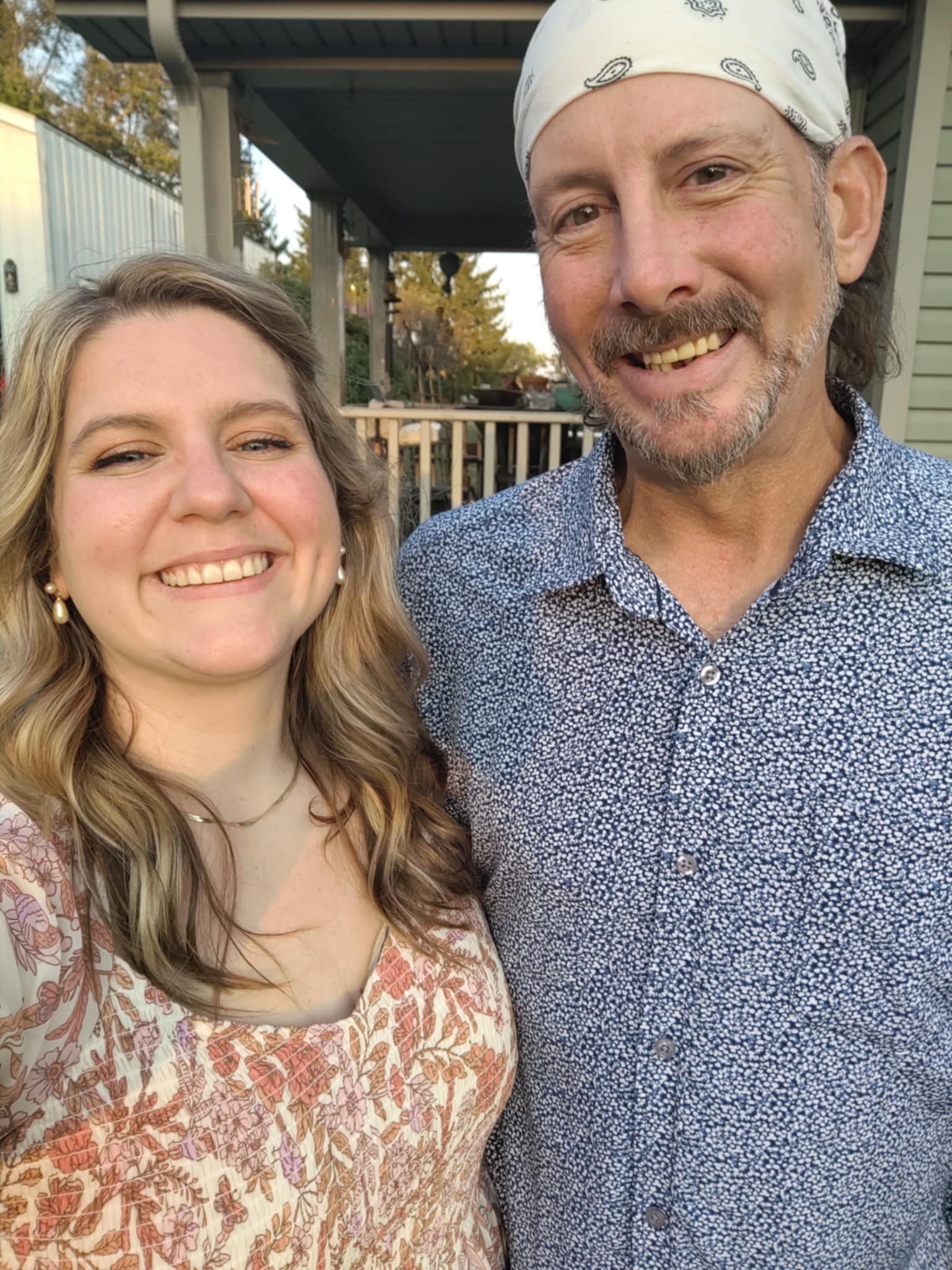 "Lauren and Tom, a joyful couple, stand close together in front of a tan-wood porch, smiling brightly for a photo. Lauren, on the left, has dirty blonde curls, bright eyes, and wears an orange and white floral top. Beside her, Tom sports a white bandana, a greying beard, and a blue button-up shirt. They lean into each other, radiating warmth, love, and the comfort of home.