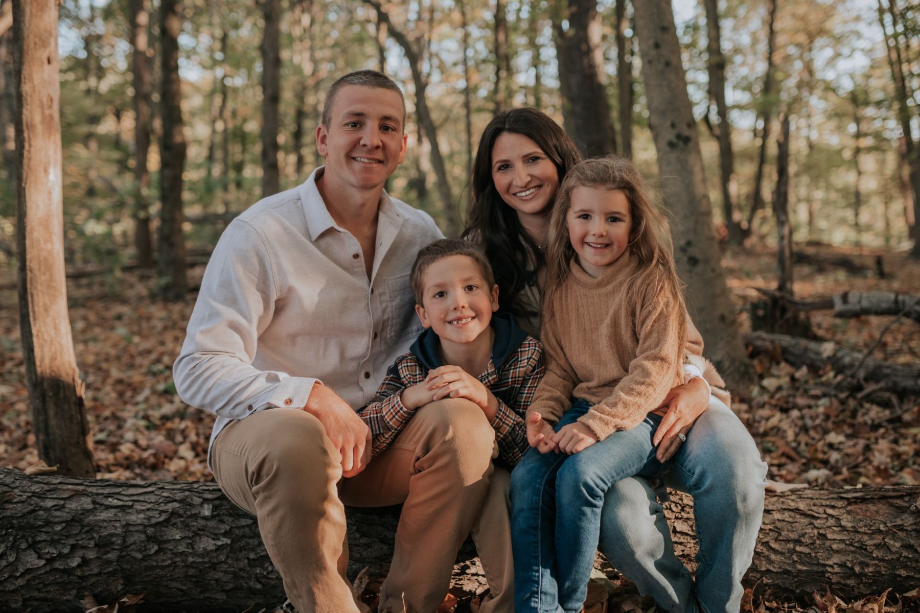 "One of our beautiful Lifespan Families, Rachel and Michael, sit with their two children on a fallen tree in a quiet, leaf-covered forest. The brown-toned color palette blends seamlessly with the autumn setting. On the left, Michael wears khaki pants and a white button-down shirt, with their young son leaning against his knee—mostly hidden, adding a candid touch. To the right, Rachel, with dark hair, holds their daughter on her lap. Both girls wear matching light blue jeans and soft tan tones, echoing the family’s coordinated, natural look. It’s a serene, aesthetic fall moment filled with warmth and togetherness.