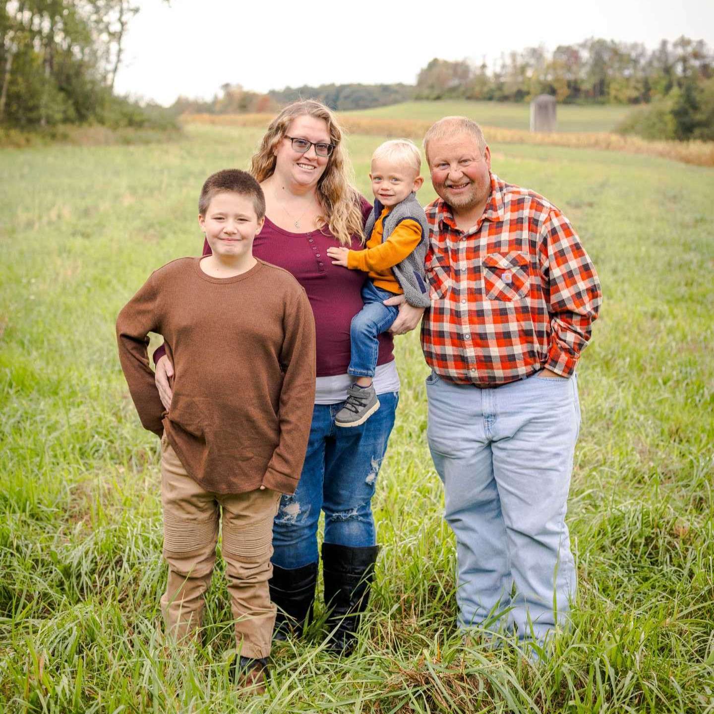 A family of four stands together in a green field, dressed in cozy, cottage-farm style outfits. The little boy, nearly as tall as his mom, stands confidently with one hand on his hip and the other relaxed by his side. He wears khaki pants and a brown top. Behind him, Mom smiles brightly, sporting dirty blonde curly hair, black glasses, tall black farm boots, blue jeans, and a burgundy top layered over grey. She holds their blonde toddler, who wears a vest, blue jeans, and an orange top, with bleach blonde hair catching the light. Dad stands nearby, smiling with a goatee, dressed in a classic red-and-white checkered farmer shirt and light blue jeans. Trees and a blue sky frame the peaceful rural scene.