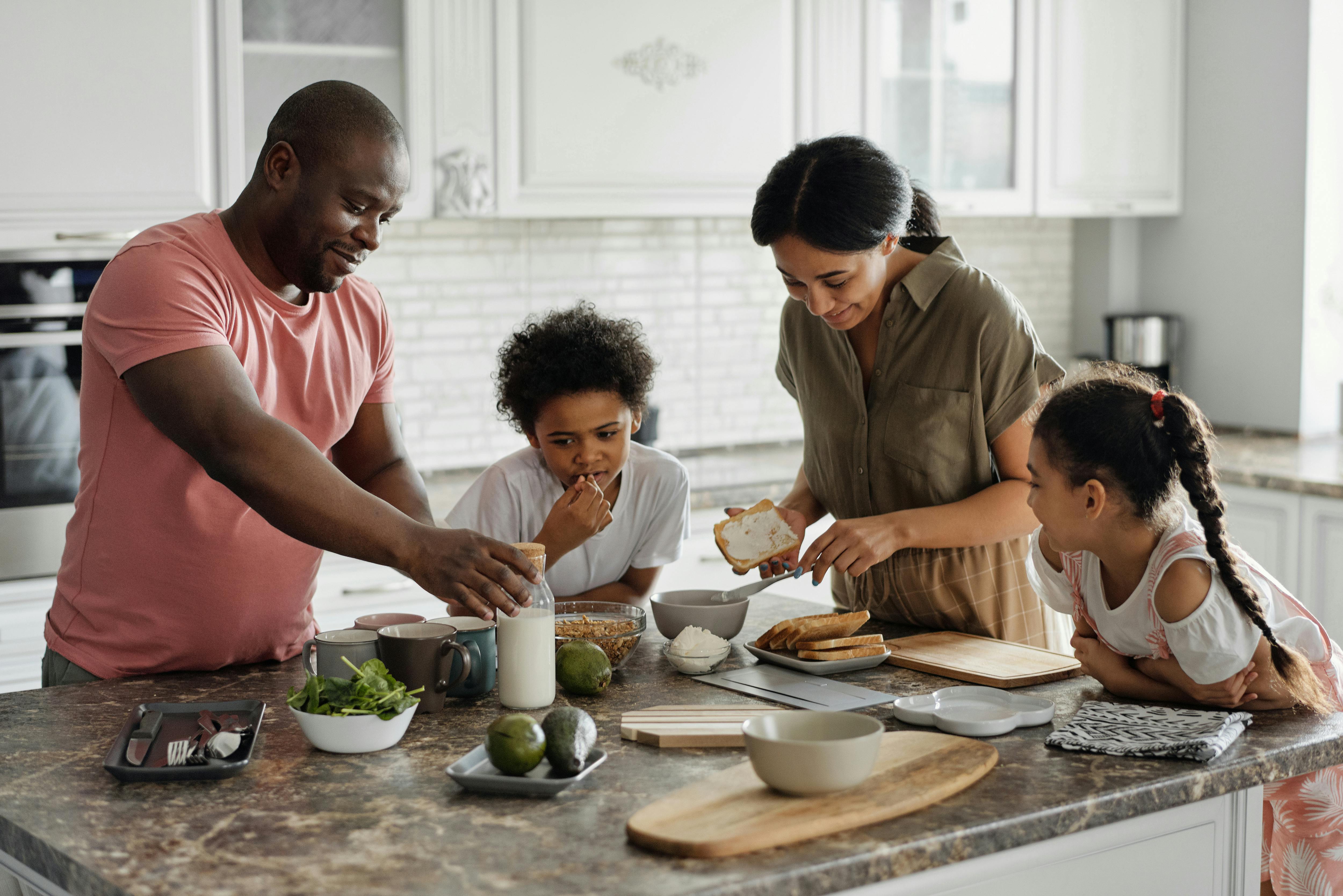 A family of four gathers in a bright, modern white kitchen, sharing a warm morning moment around a kitchen island. A Black adult male in a peach shirt leans in beside a young Black boy, both smiling. Across from them, a darker-skinned adult woman and a little girl with matching features lean in close, likely enjoying breakfast together. The scene radiates togetherness, comfort, and the simple joy of family connection.
