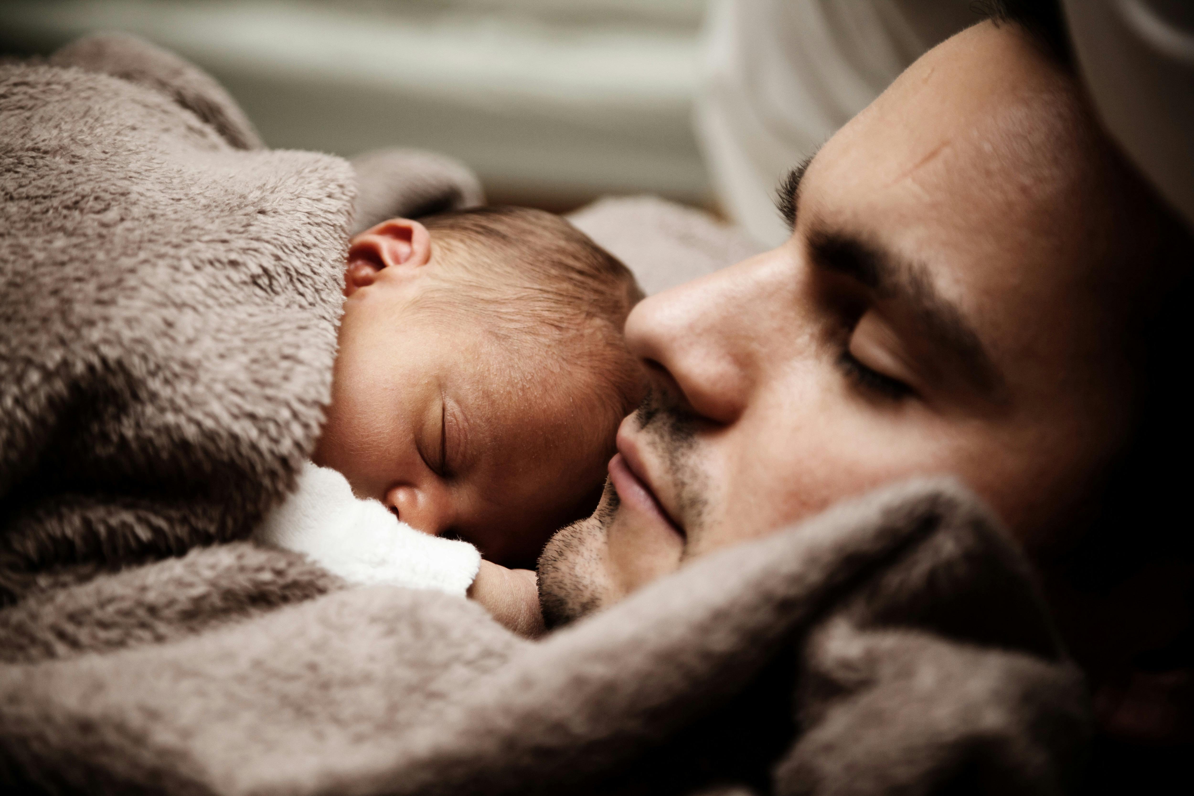 A man gently cradles a newborn baby against his chest, eyes closed in a quiet, emotional moment of connection. The baby is nestled into a soft, fleecy wool blanket, creating a sense of warmth, safety, and tender bonding between them.