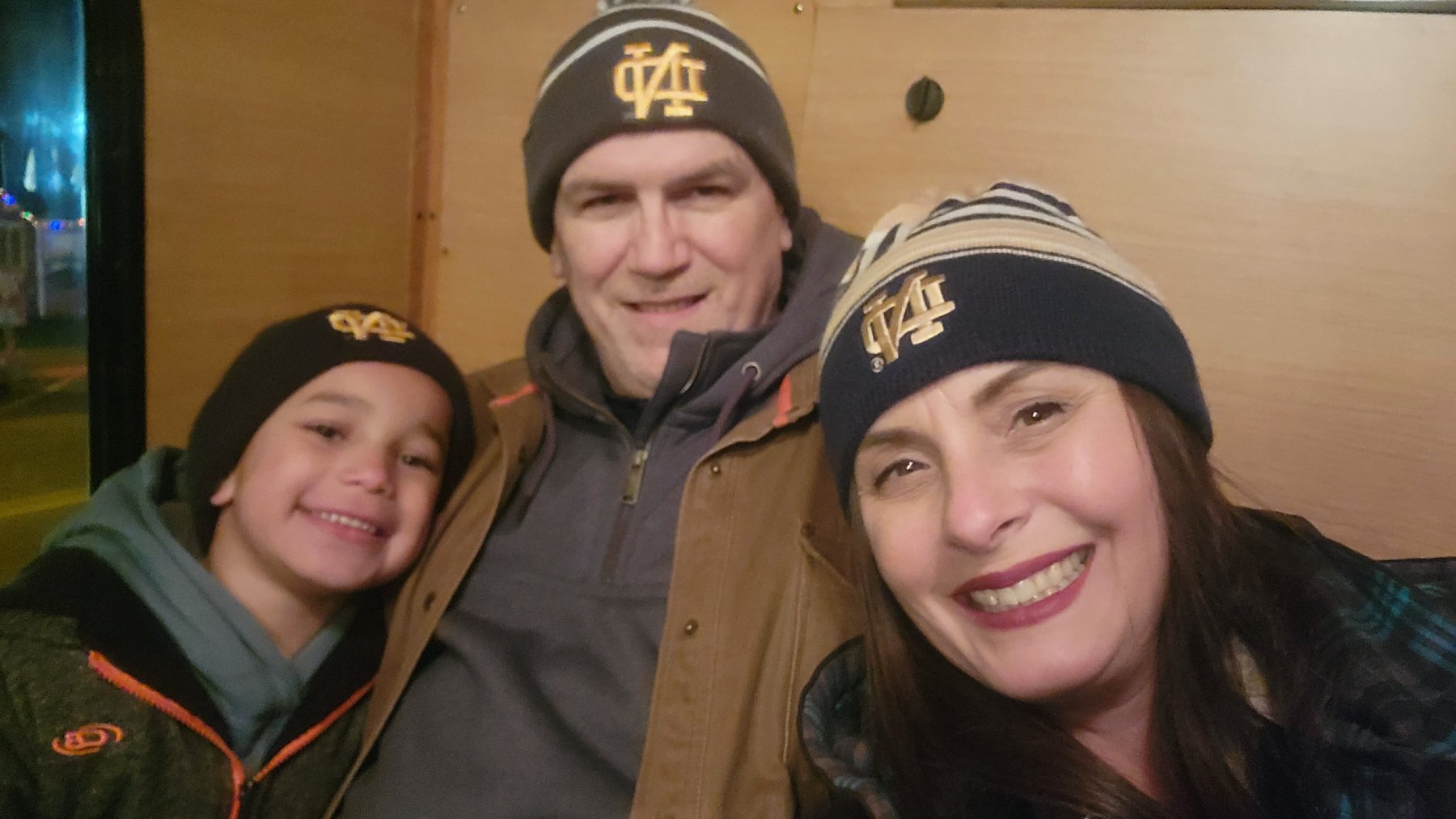 "One of our Lifespan Families—Jeff, Rhonda, and their son—pose closely together for a cheerful selfie. The little boy leans into his dad, Jeff, while Rhonda leans in from the right with a warm smile. All three wear matching hats and cozy layered clothing, against a tan wall. The little boy beams brightly, and Rhonda’s dark hair and bold red lipstick add a pop of color to the cozy, winter-ready look. The boys' hair is hidden beneath their hats.