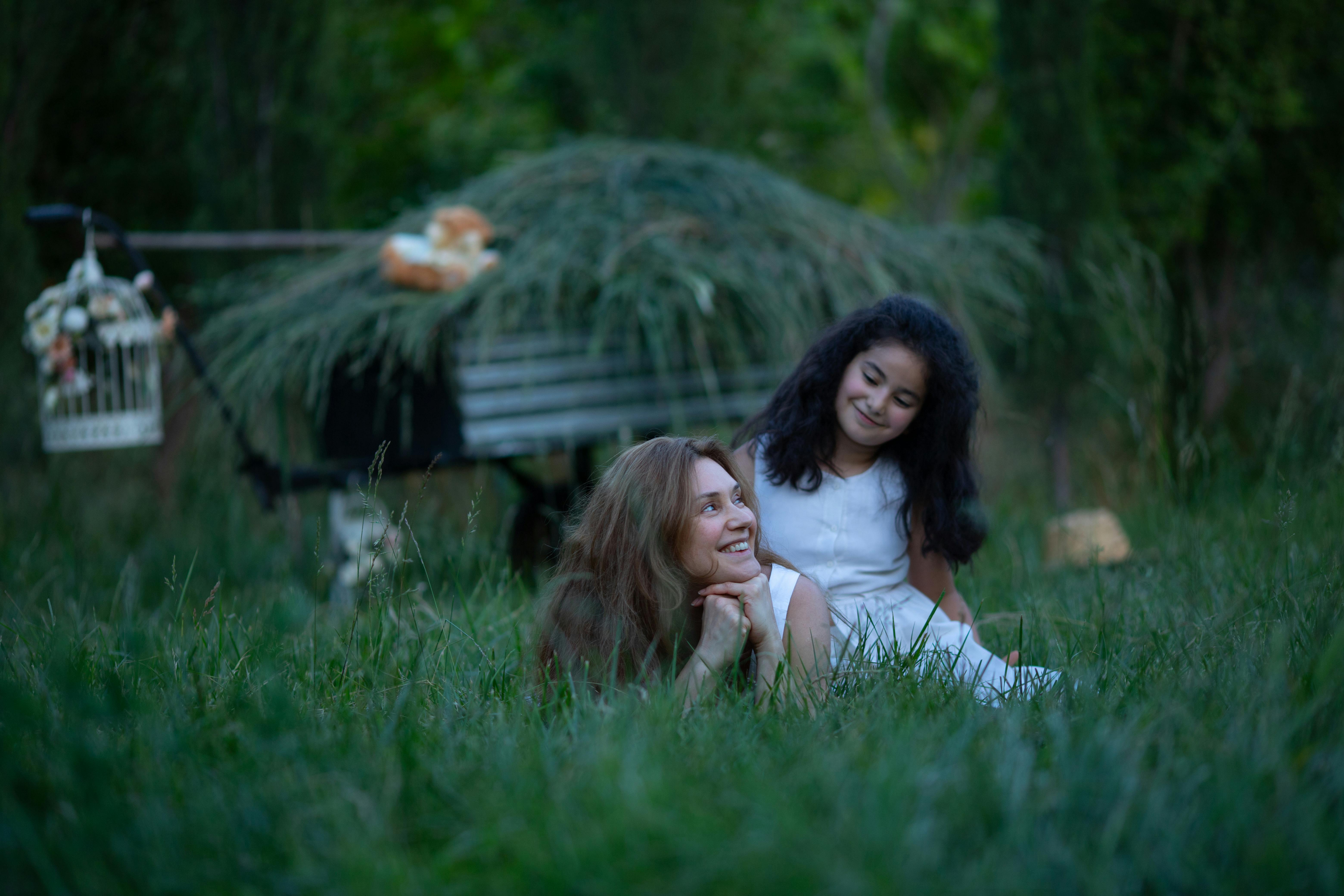 "A young girl with black hair and an older woman share a joyful smile as they face each other in a sunlit grassy field. The moment captures warmth, connection, and the bond between generations in a peaceful outdoor setting.