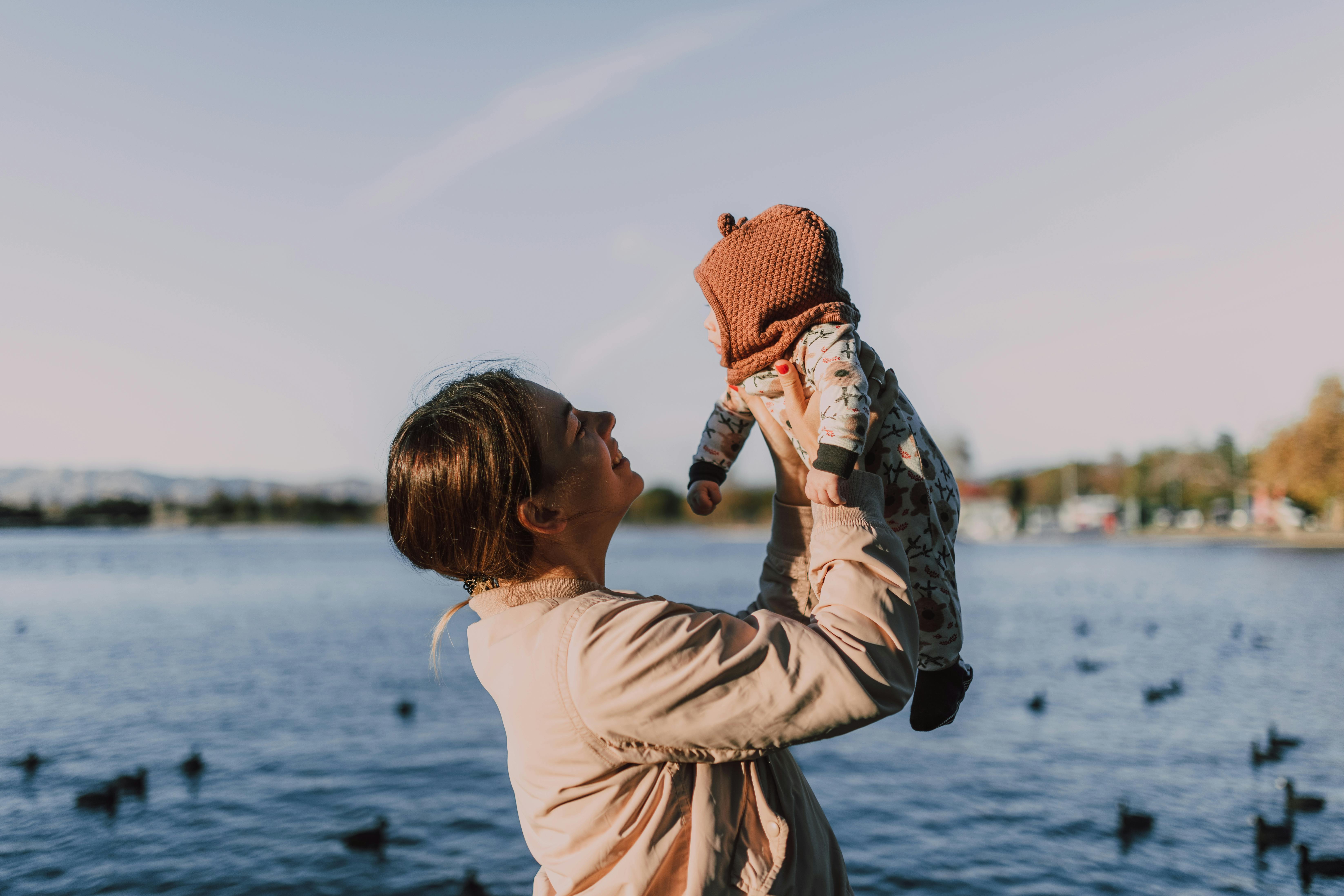 A photo of a woman holding a baby up. She smiles at the baby with adoration. They stand in front of a body of water on a cloudy, gray day.