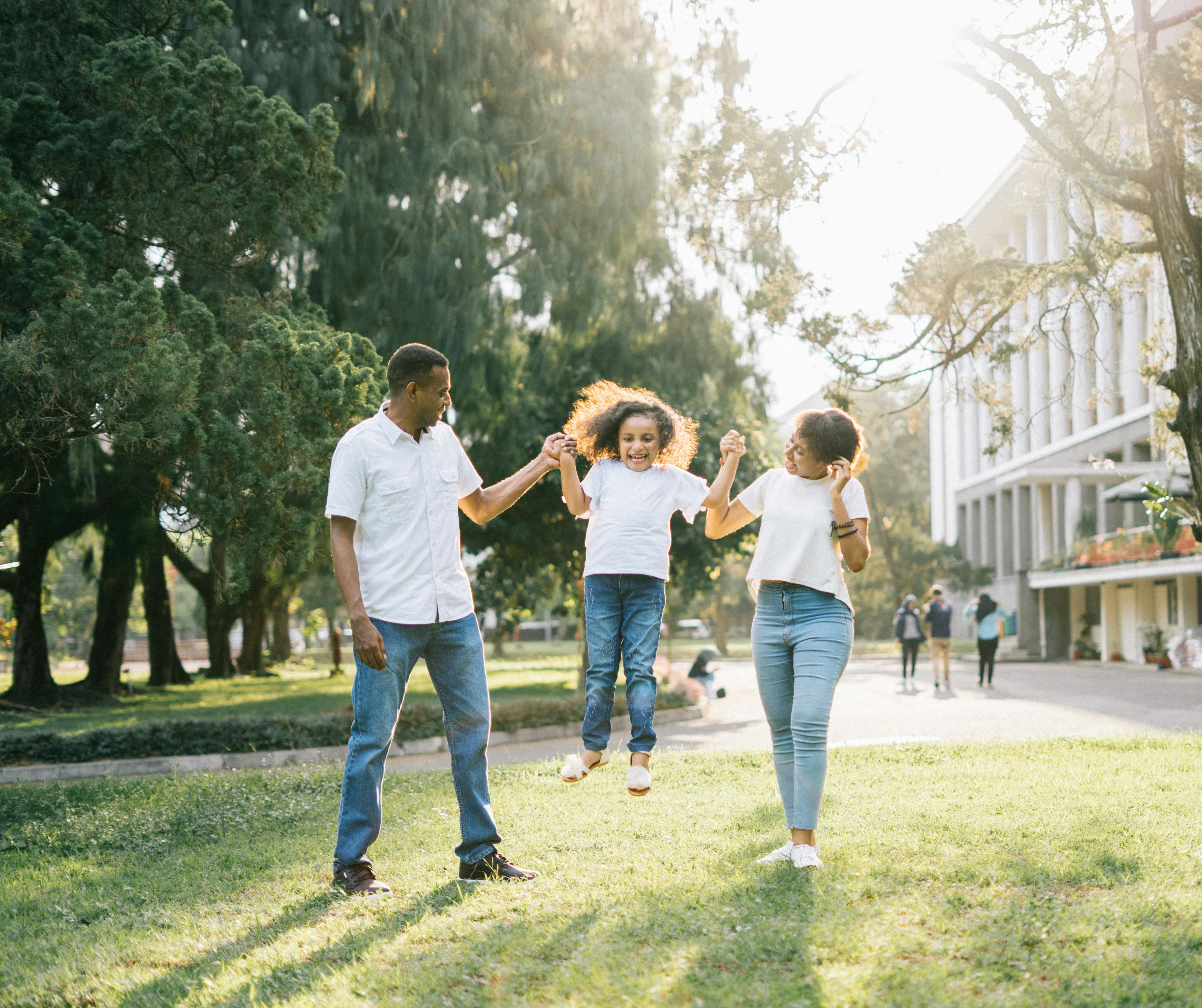 Two parents hold the hands of a child as she jumps for joy into the air. They are in the middle of a green area, with a sidewalk and buildings behind them. They all are dressed in white t-shirts & blue jeans. 