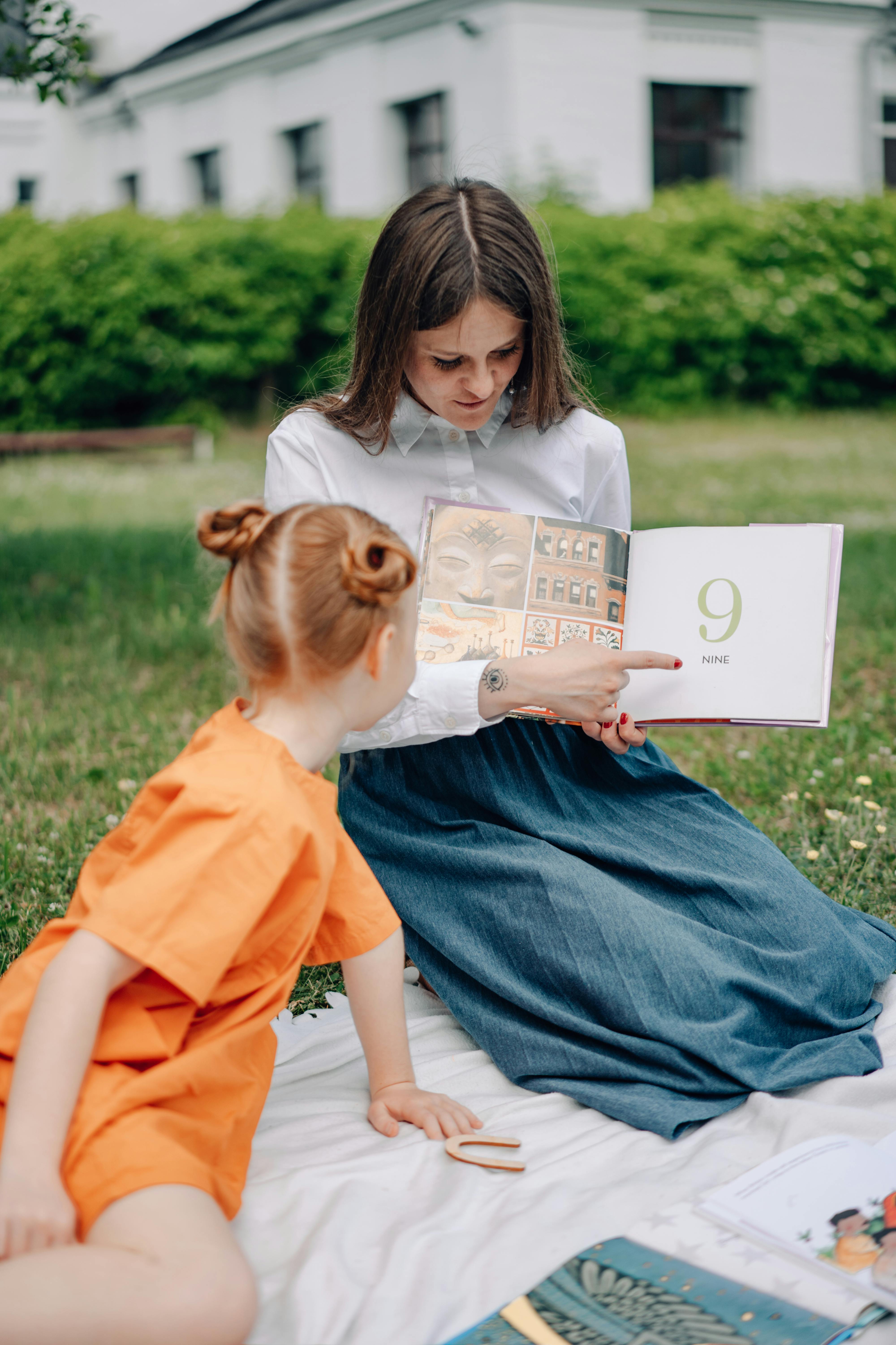 A woman in a dress shows a book to a little girl. 
