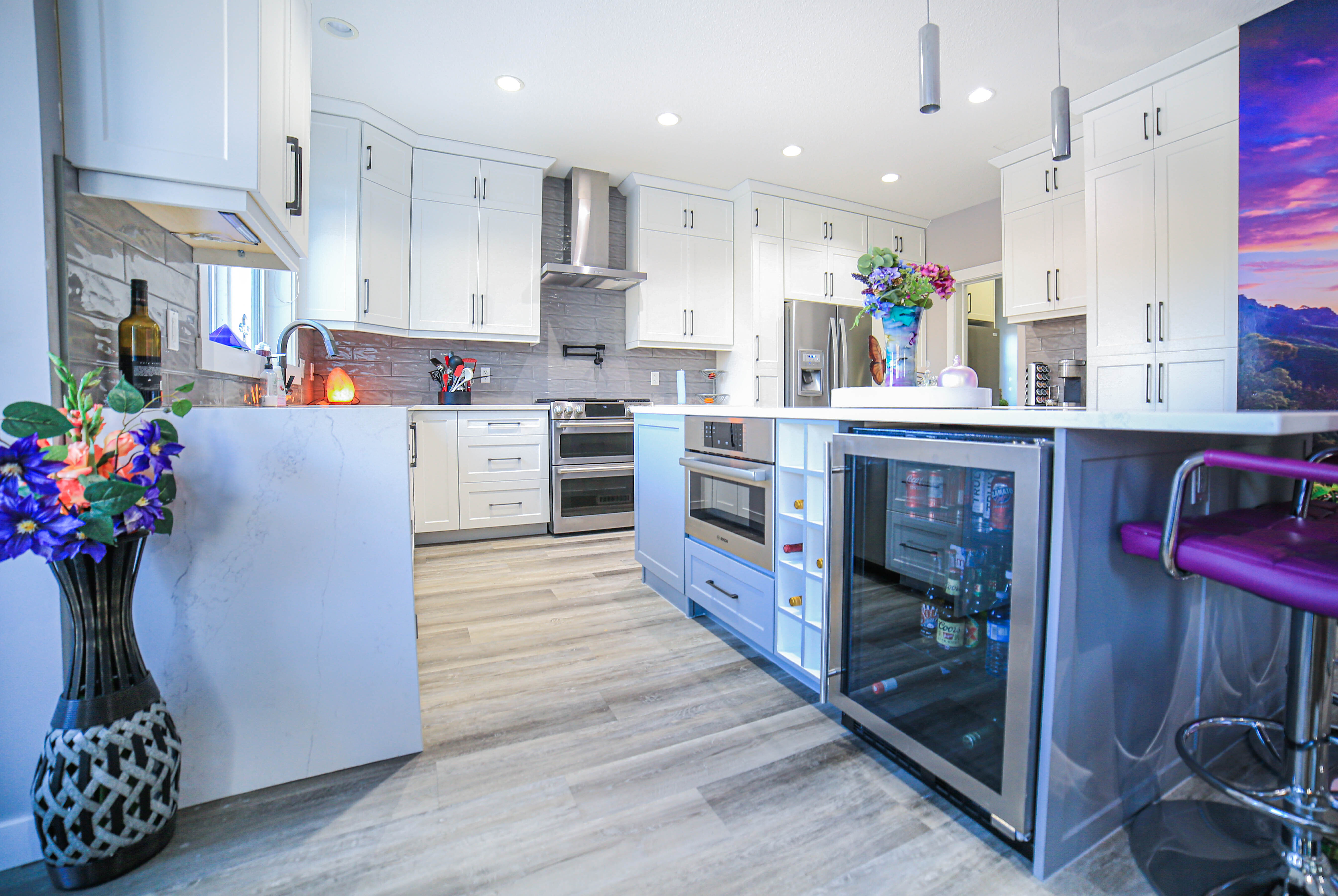 A modern, bright kitchen featuring white shaker cabinets, a grey island with a built-in wine fridge, stainless steel appliances, and light wood-look flooring.