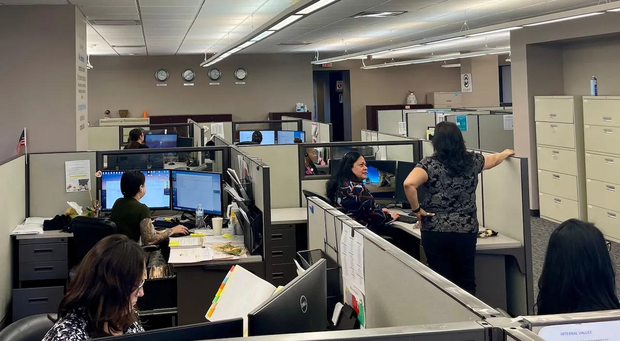 Office workspace with multiple employees working at cubicles equipped with computers and office supplies.