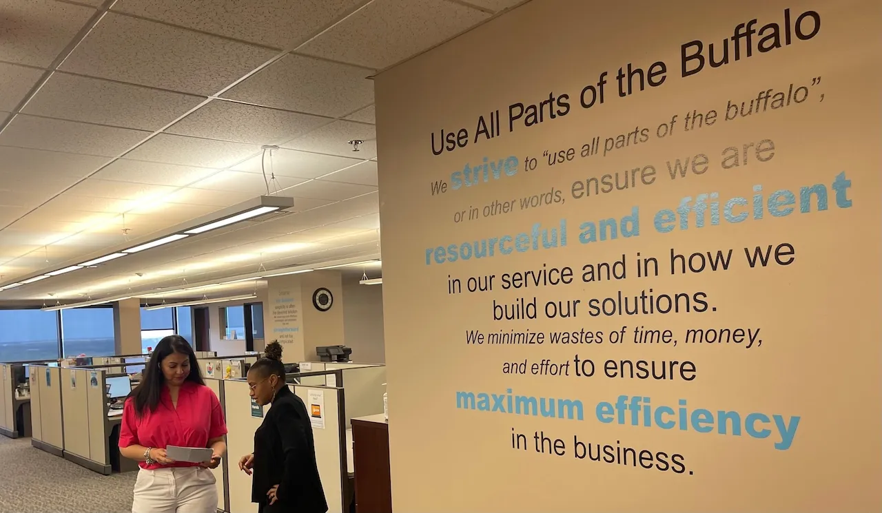 Two women standing in an office hallway near a wall displaying the text 'Use All Parts of the Buffalo' with a message about striving to be resourceful and efficient in service and solutions.