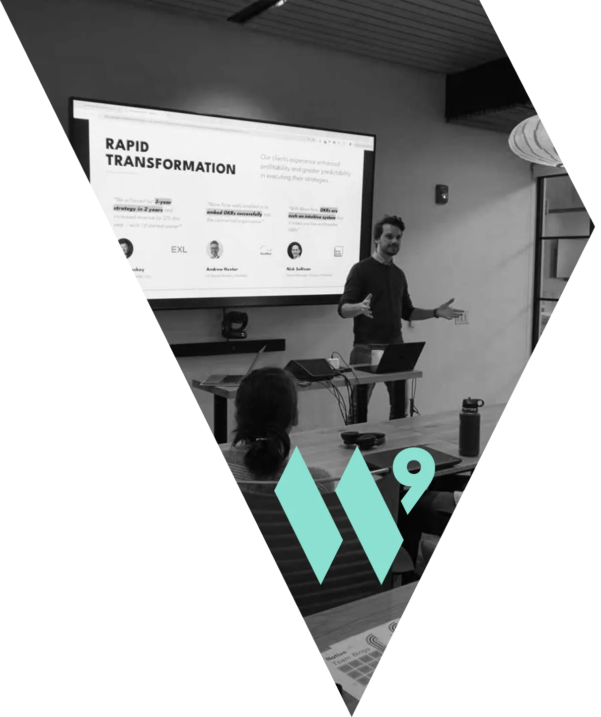 A man stands at the head of a long wooden table in a contemporary meeting room, actively delivering a presentation. The presentation, titled "RAPID TRANSFORMATION," is displayed on a large screen behind him and discusses OKR (Objectives and Key Results) implementation. The image is shot in black and white, creating a professional and focused atmosphere. Seated at the table, a woman is visible from behind. In the foreground, the vibrant teal logo of the company, WaveNine—a stylized 'W'—is prominently overlaid on the scene. The entire composition is framed within a sharp, angular diamond shape.