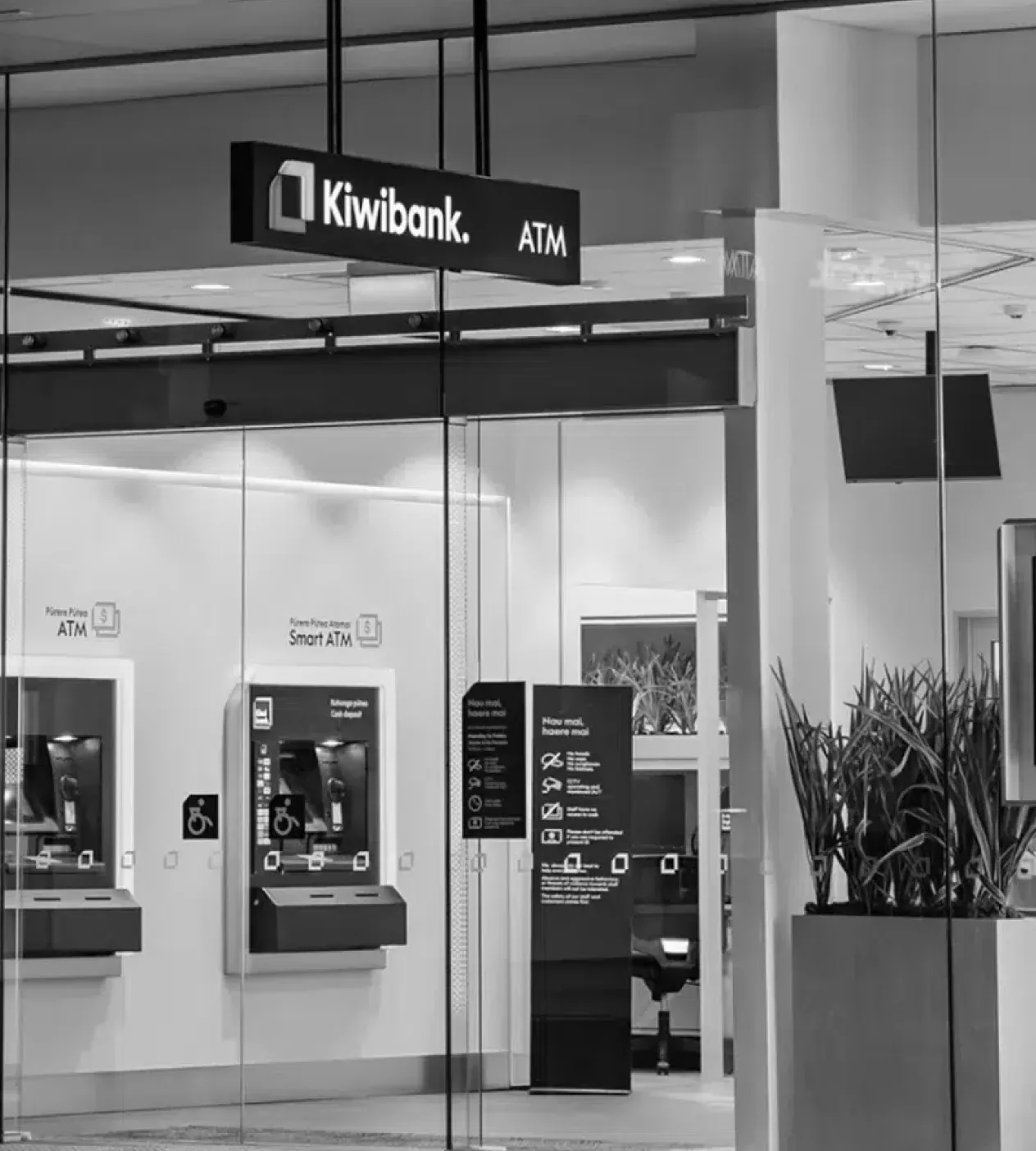 This is a clean, black and white shot of the entrance to a Kiwibank ATM area, likely within a larger building or branch. A prominent sign hangs from the ceiling, reading "Kiwibank. ATM" with the bank's logo. Through the glass walls and entrance, two ATMs are mounted on the far wall; one is labeled "ATM" and the other "Smart ATM." Both have accessibility symbols. To the right, a large planter with a leafy plant adds a touch of green, though rendered in grayscale. The overall aesthetic is modern and professional.

