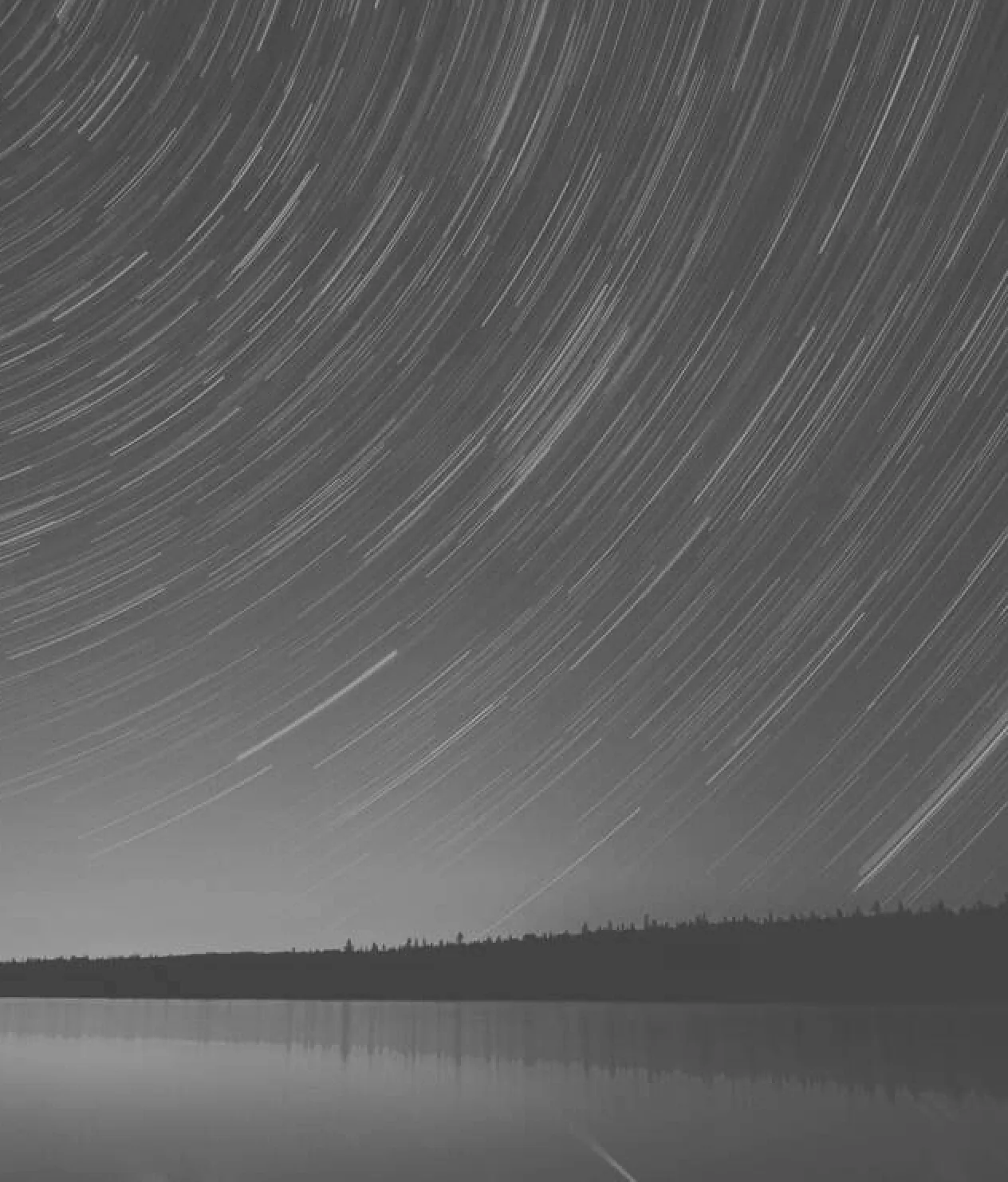 This is a serene, black and white, long-exposure photograph of a night sky over a calm landscape. The sky dominates the frame, filled with countless streaks of light which are trails left by stars, illustrating the rotation of the Earth. These trails arc and sweep diagonally across the sky. Below, a dark, silhouetted treeline sits on the distant horizon. The foreground is a smooth, glass-like body of water that mirrors the bright star trails from the sky above, creating a sense of symmetry and tranquility.