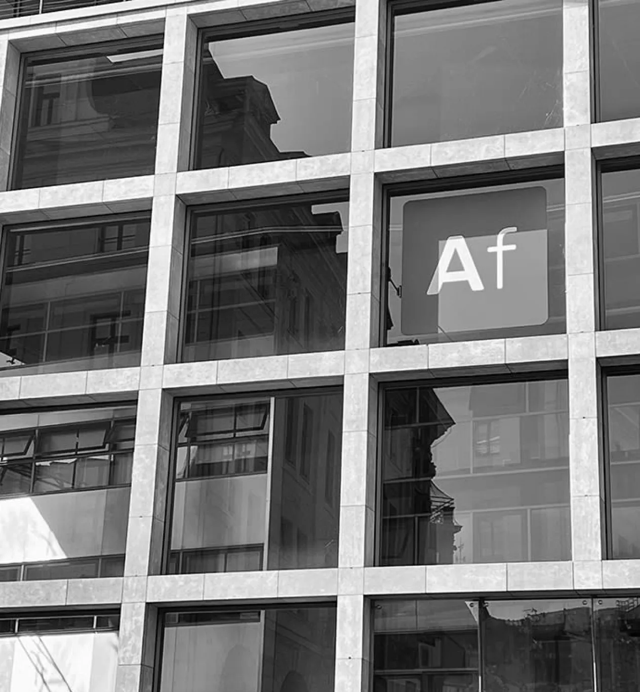 This is a black and white, angled shot of a modern building's facade, characterized by a strong grid pattern of concrete or stone. The grid frames large, square windows that reflect the distorted images of older, more ornate buildings from across the street, creating a contrast between architectural styles. In one of the upper-right windows, a lit, square sign with rounded corners is visible, featuring the white letters "Af" in a clean, sans-serif font, similar in style to an Adobe Creative Cloud application logo. The monochromatic tones of the photo emphasize the building's textures, lines, and geometric structure.