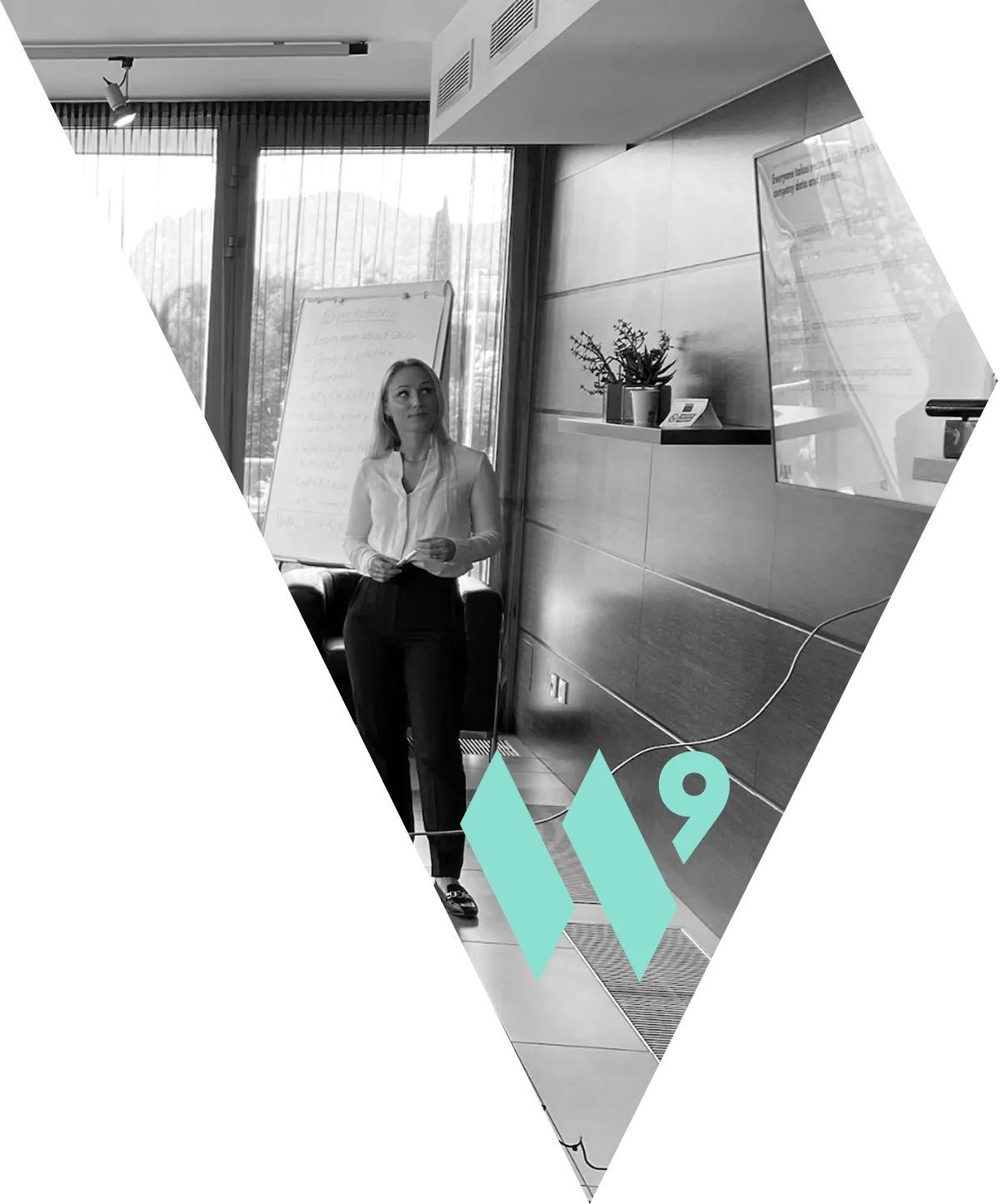 This image captures a woman in professional attire (a white blouse and dark trousers) standing confidently in a modern meeting room. The room is filled with natural light from a large window that reveals a mountain in the background. To her side stands a flip chart with handwritten notes, suggesting a presentation or collaborative session. The photo is presented in black and white and is framed within a pointed, diamond-like shape against a black background. Overlaid at the bottom is the prominent, colorful teal logo for the company WaveNine.