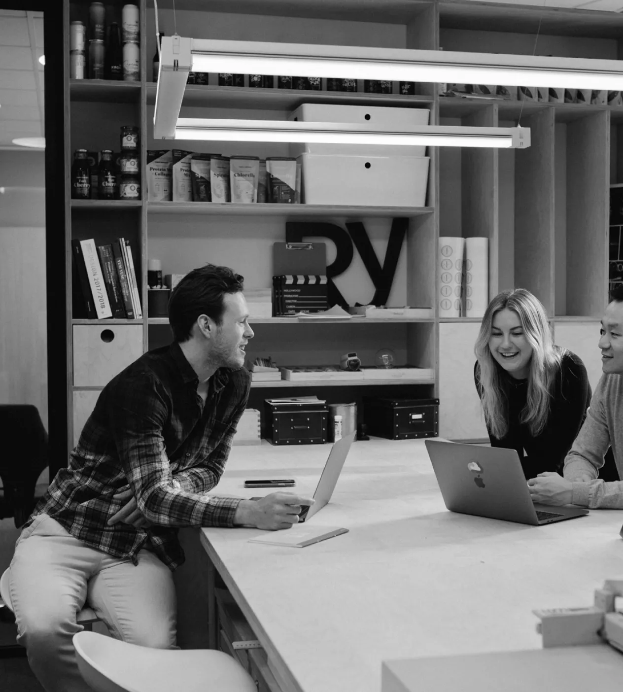 This black and white photo captures a positive and collaborative atmosphere in a modern office. Three colleagues are gathered around a large, light-colored wooden table, each with a laptop. A man in a plaid shirt on the left leans forward on a stool, smiling and engaging with a woman in the center, who is also smiling brightly. A third colleague is visible on the right. The setting is a stylish creative space, with large built-in shelves in the background holding books (one on "BRANDING"), decorative items like a film clapperboard, and large letters spelling "RV". A sleek, modern light fixture illuminates the scene.