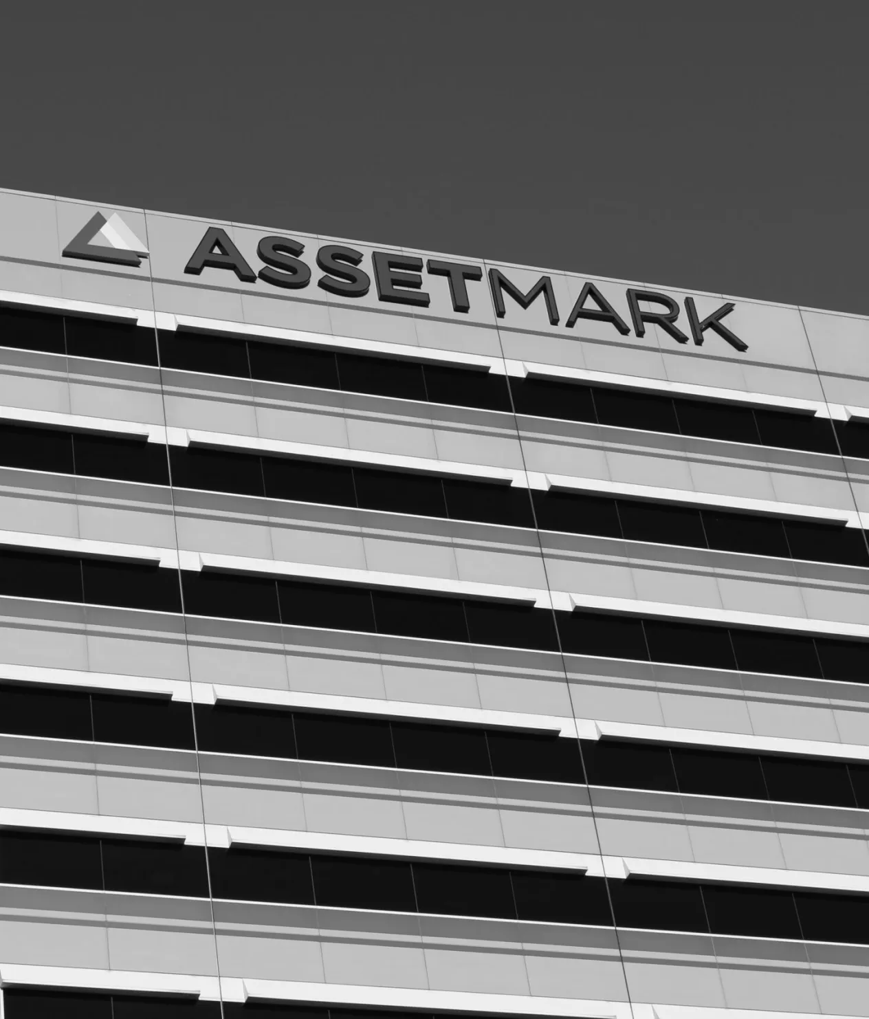 A low-angle, black and white photograph of the AssetMark logo and sign on the facade of a modern office building.