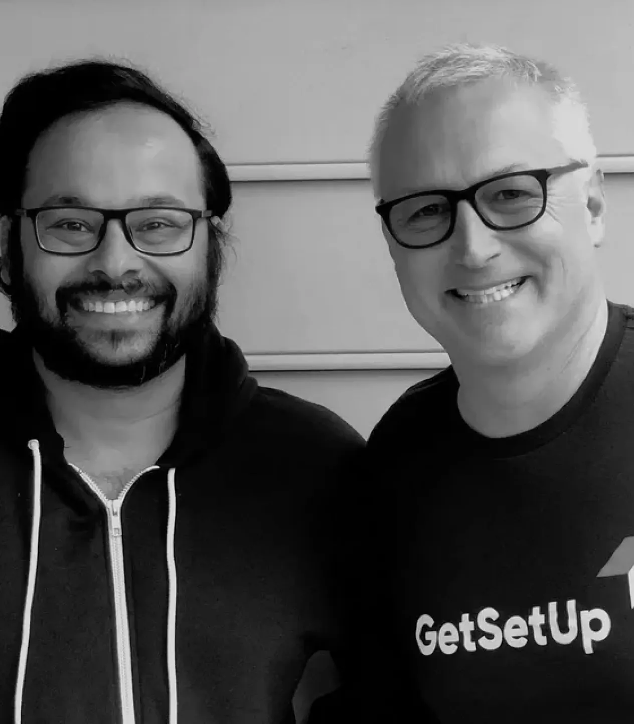 A black and white photo of two men standing side-by-side and smiling at the camera. The man on the right is wearing a t-shirt with the "GetSetUp" logo.