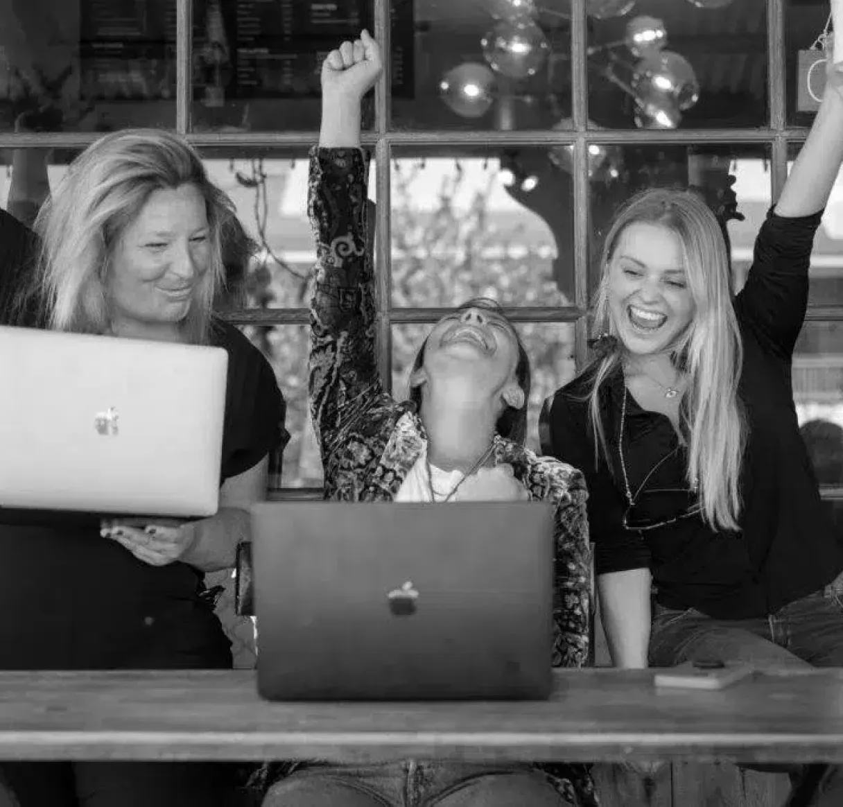 A black and white photo of three women at a table with laptops, celebrating a major success. Two of the women have their arms raised in excitement, illustrating a moment of team achievement and collaboration.