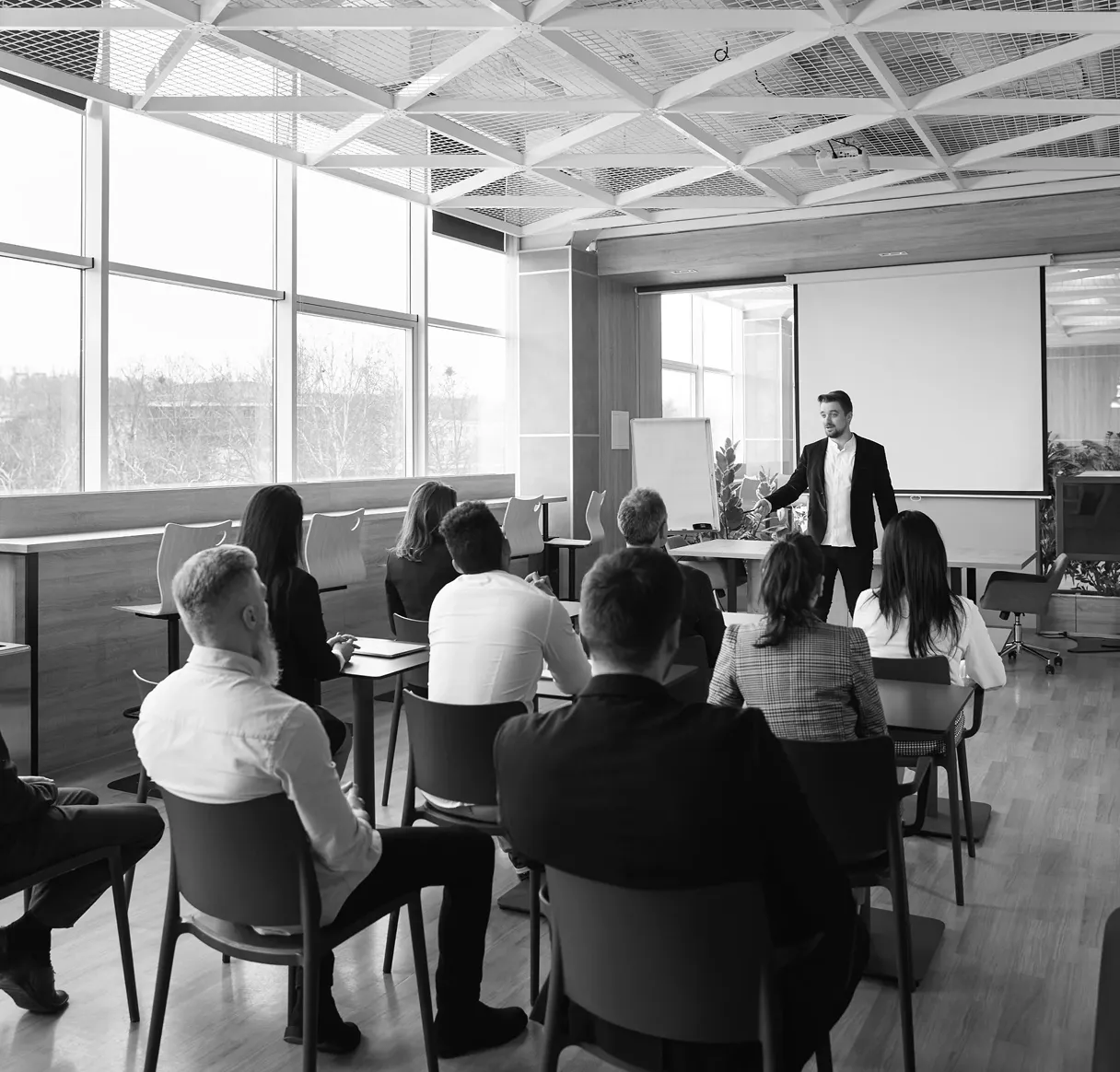 A black and white photo showing a business training session in progress. A male presenter from Wavenine stands at the front of a room with large windows, addressing a seated audience during a workshop or seminar.