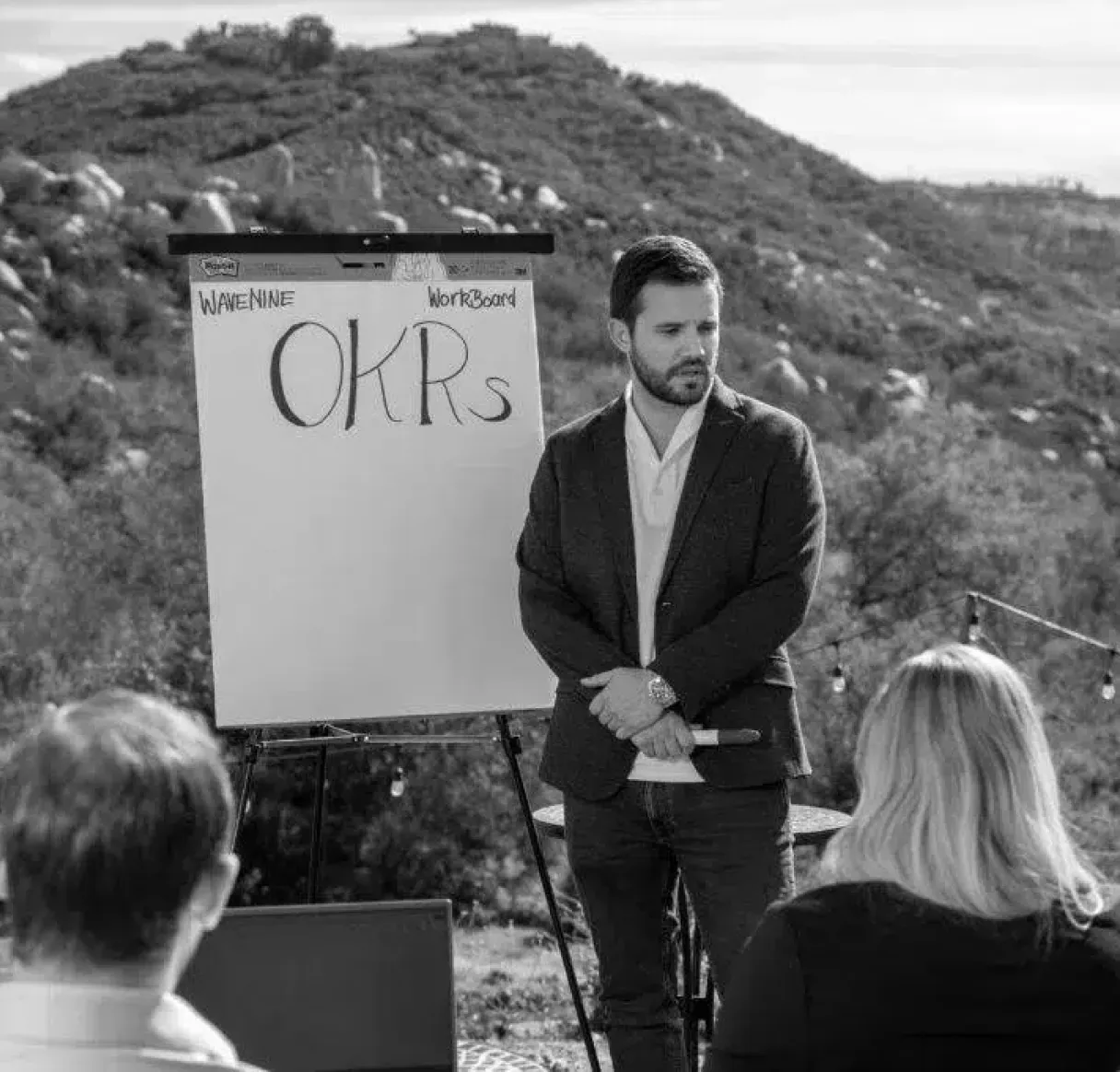 A black and white photo of a business coach from Wavenine leading a training session on OKRs in an outdoor setting. He is presenting to a small group with a whiteboard that displays the words "Wavenine," "WorkBoard," and "OKRs."