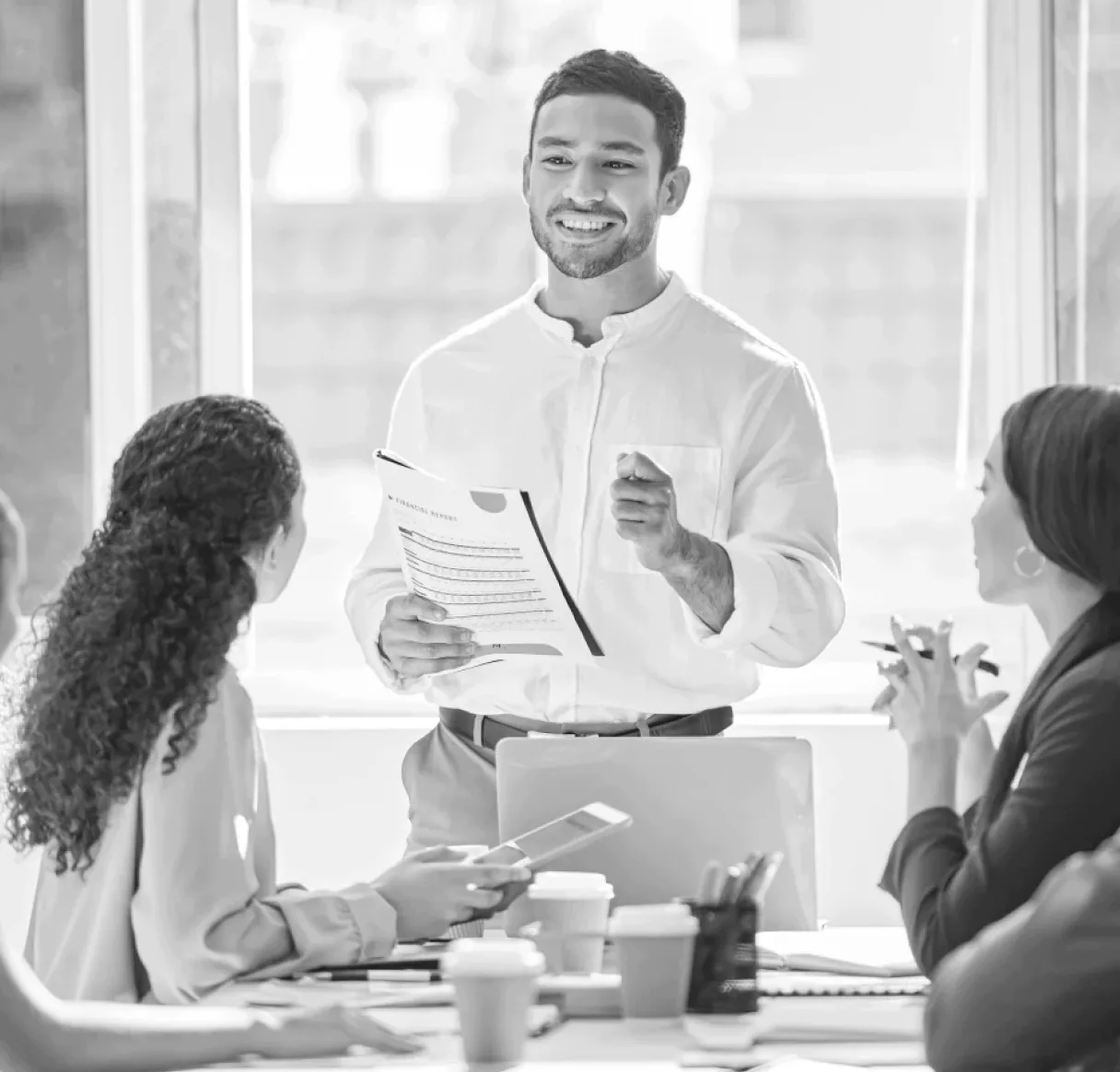 A black and white photo of a smiling manager leading a collaborative team meeting. He stands and holds a report while discussing successful performance outcomes with his team in a modern, brightly-lit office.